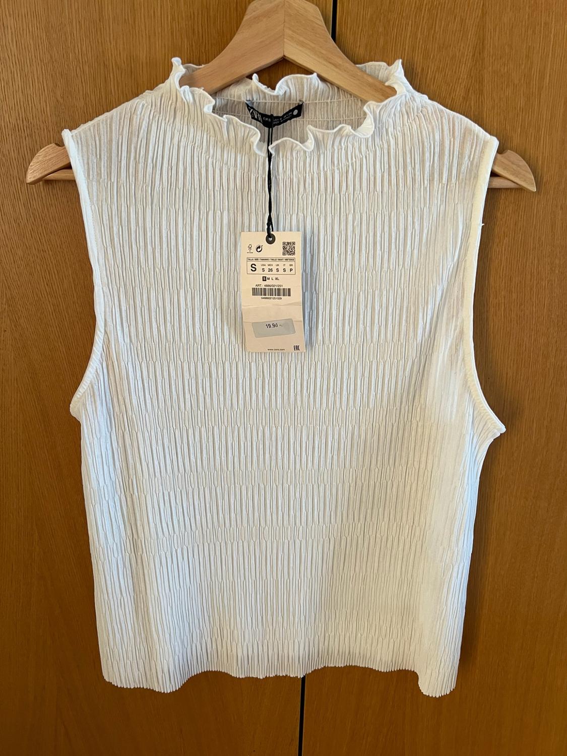 White sleeveless top, textured fabric and high ruffled neck, on a hanger against a wooden background. Text: ZARA, S, 19.90.