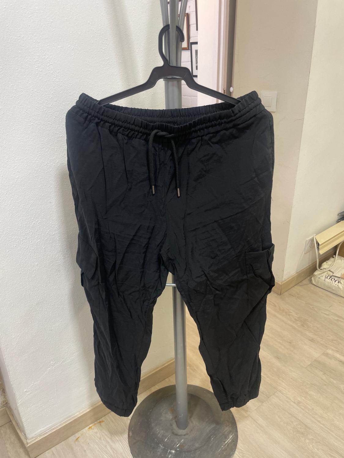 Wrinkled black trousers hanging on a hanger in an indoor setting with light wood flooring and a white wall.