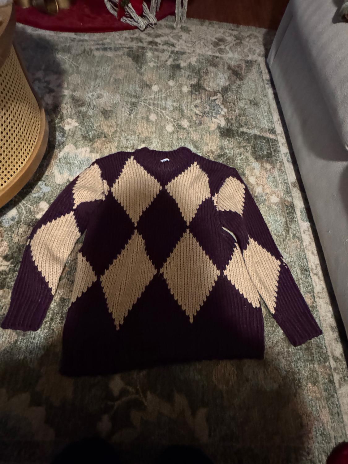 Beige and dark purple argyle jumper on a rug with a floral design in green and beige tones, next to a wicker furniture piece.