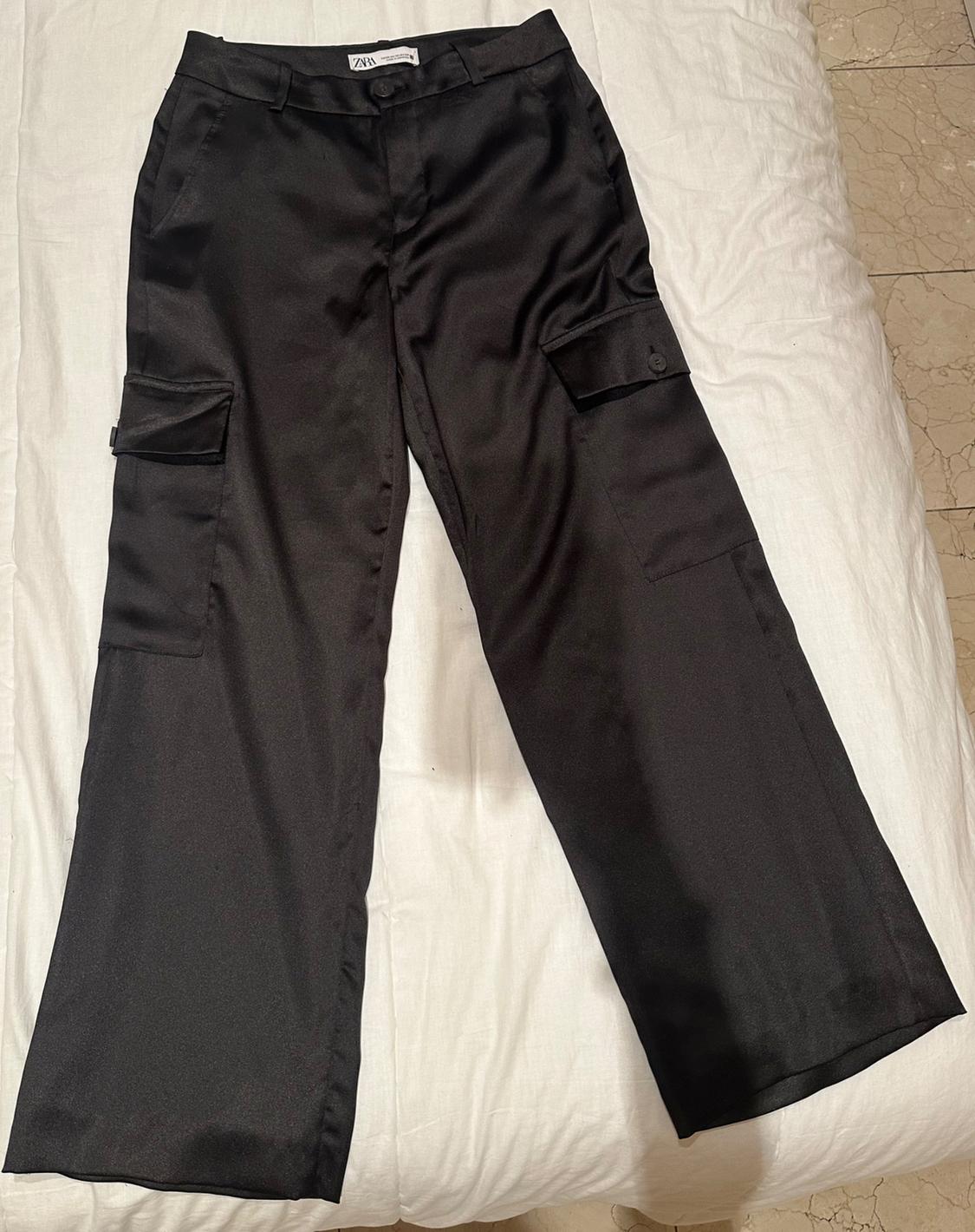 Black satin trousers with cargo pockets, placed on a white bed. Label visible on the waistband.