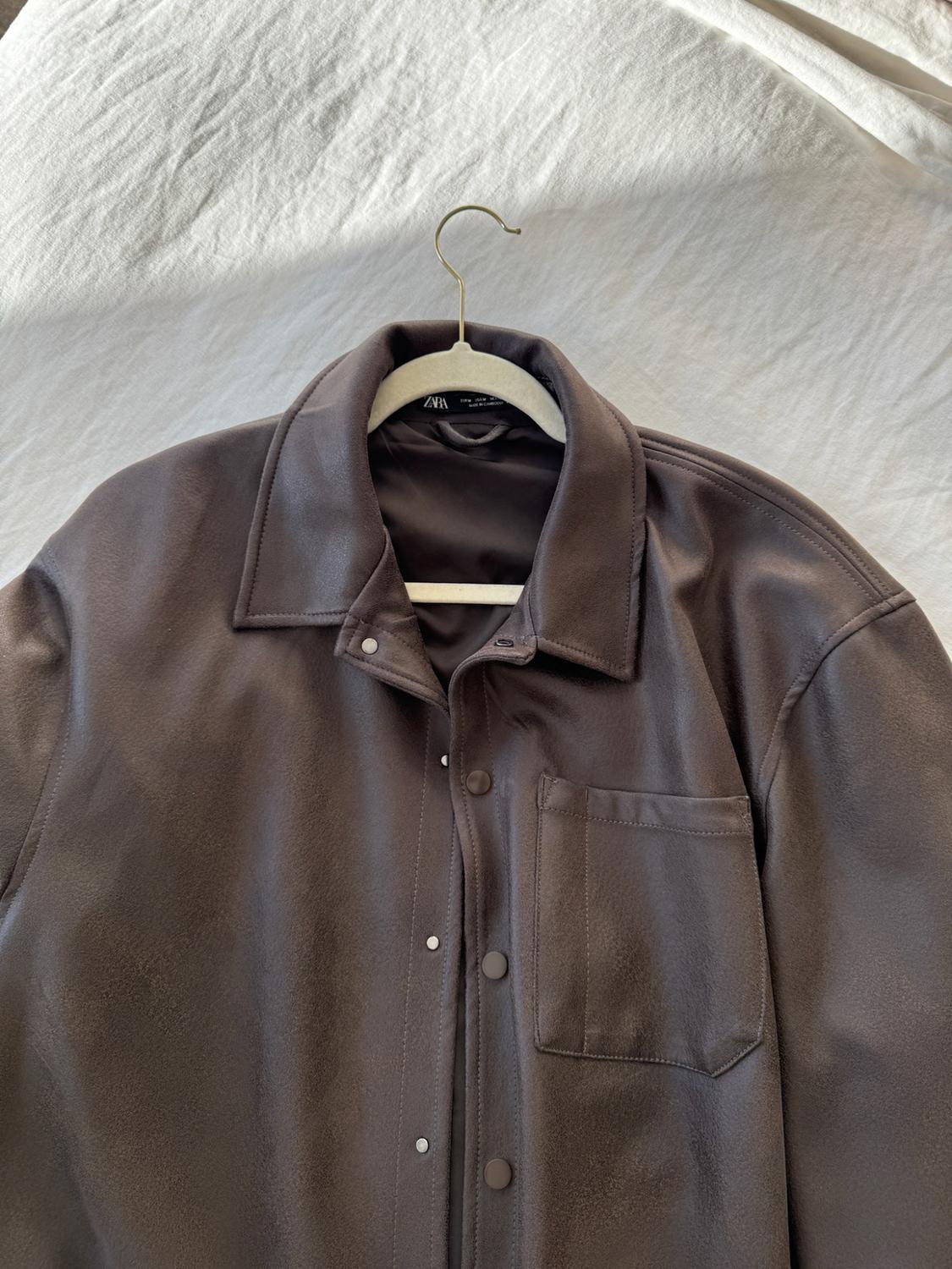 Dark brown jacket with a shirt-style collar, snap-button closure, and a front pocket, hanging on a hanger against a white background.