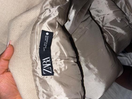Label from a Zara garment made of beige satin fabric, held by a hand. Visible text: "ZARA", "EUR L USA L MEX 30", "MADE IN MOROCCO".