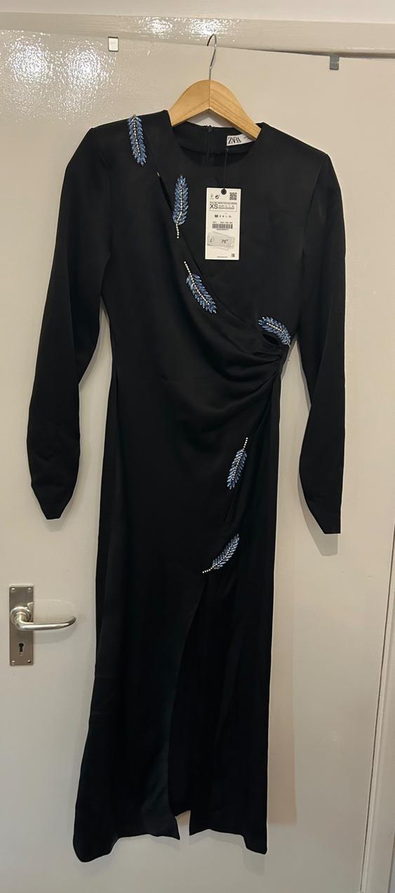 Long black dress with long sleeves, embroidered blue leaf details, hanging on a wooden hanger over a white door.