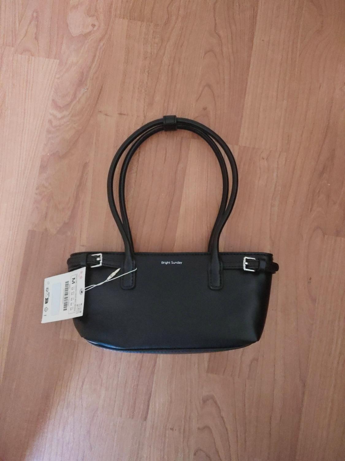 Black handbag with two handles, silver buckles and a tag on a wooden floor. Visible text: Bright Sunday.