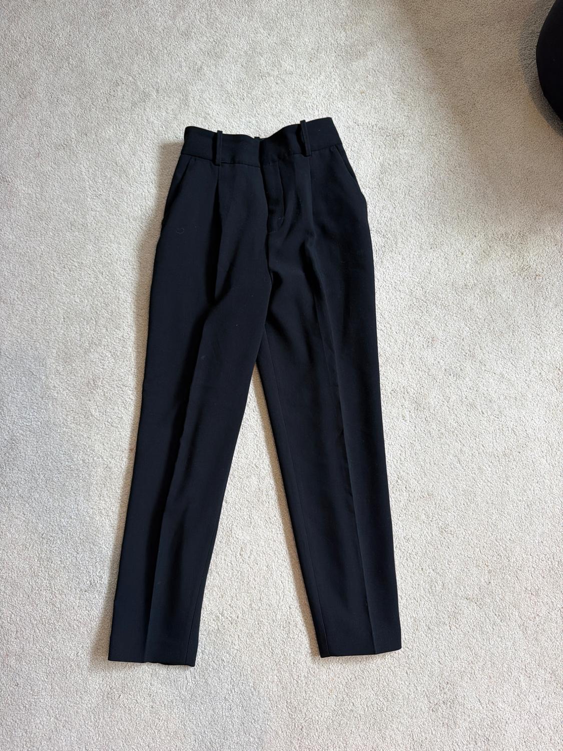 Black tailored trousers with pleats, photographed from above on a light-coloured rug in a relaxed setting.