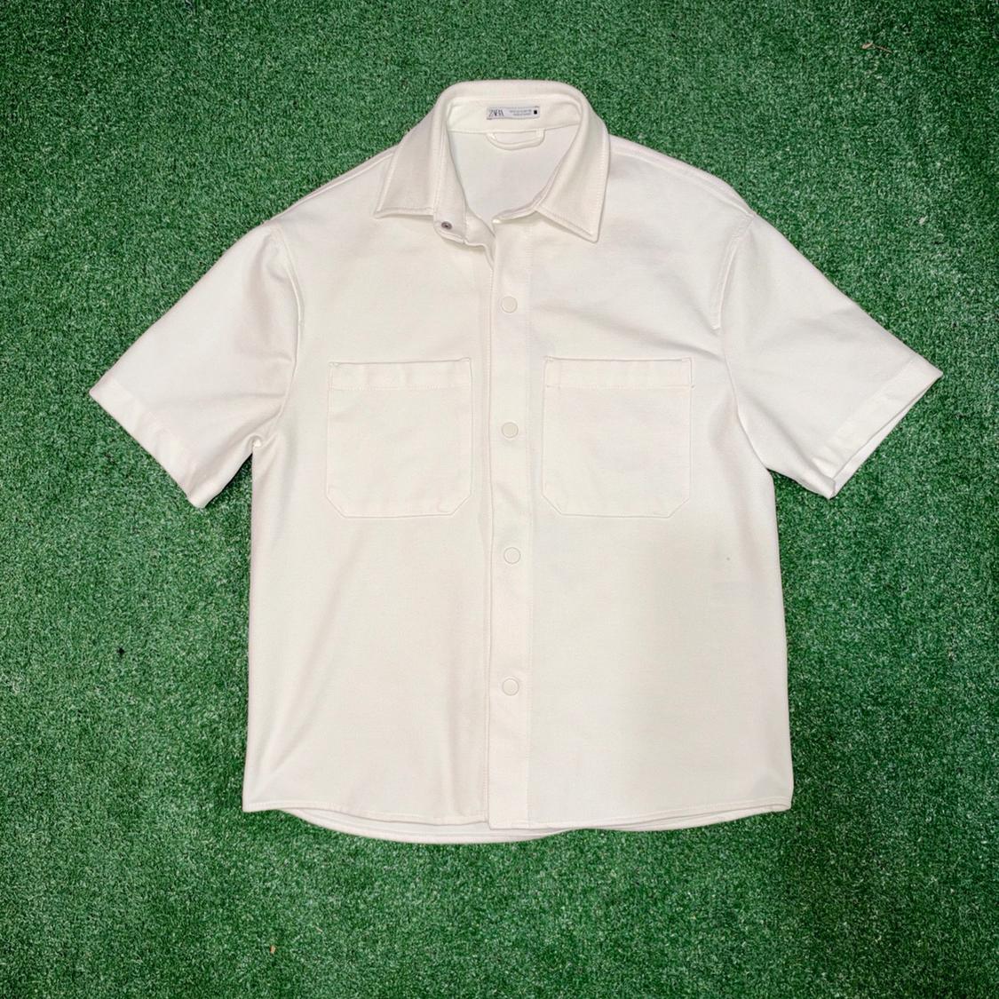 White short-sleeved button-down shirt with two front pockets, on a green artificial turf background.