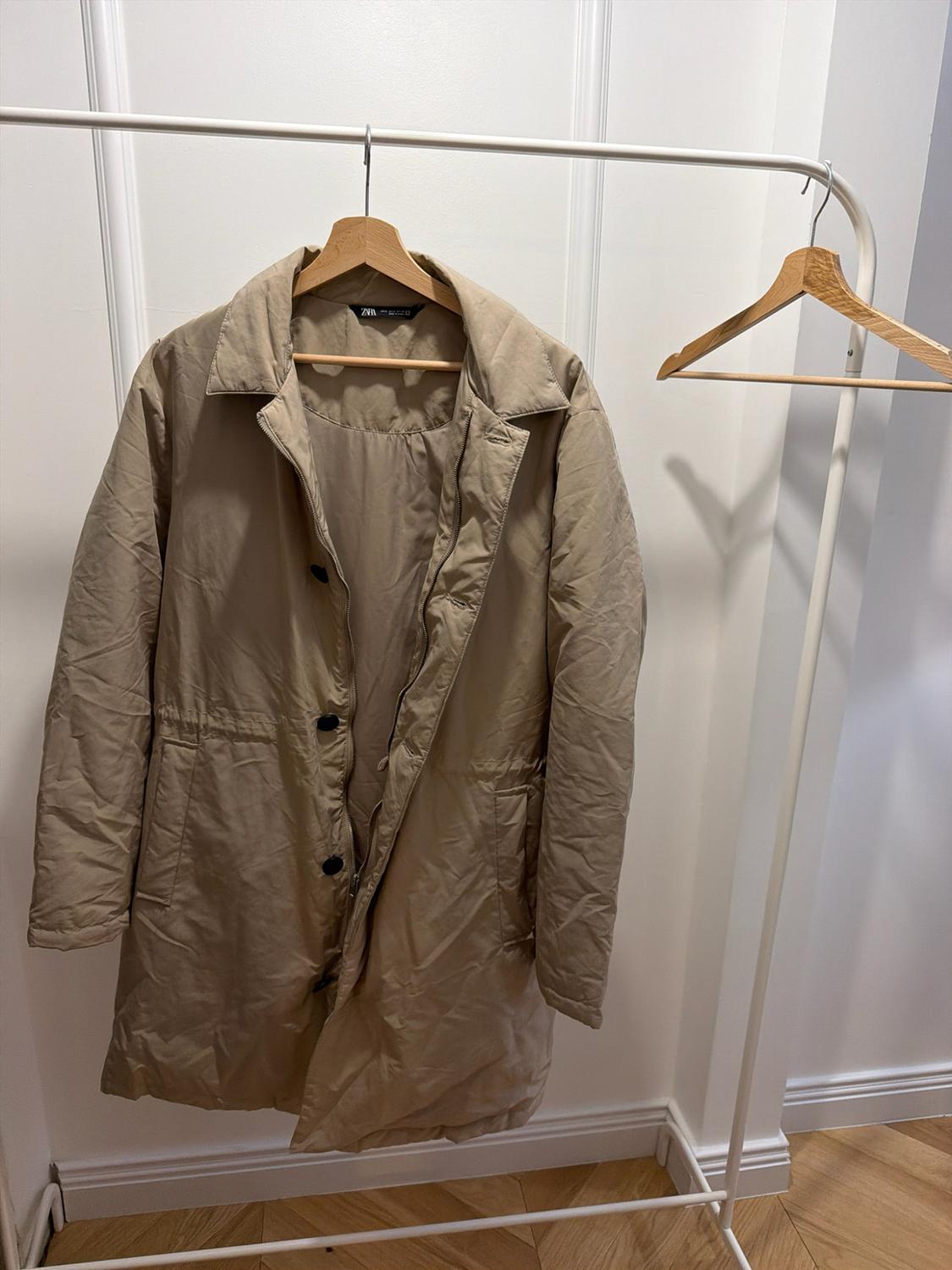 Beige coat on a wooden hanger on a white coat rack. White wall background and parquet floor. Interior label with the text ZARA.