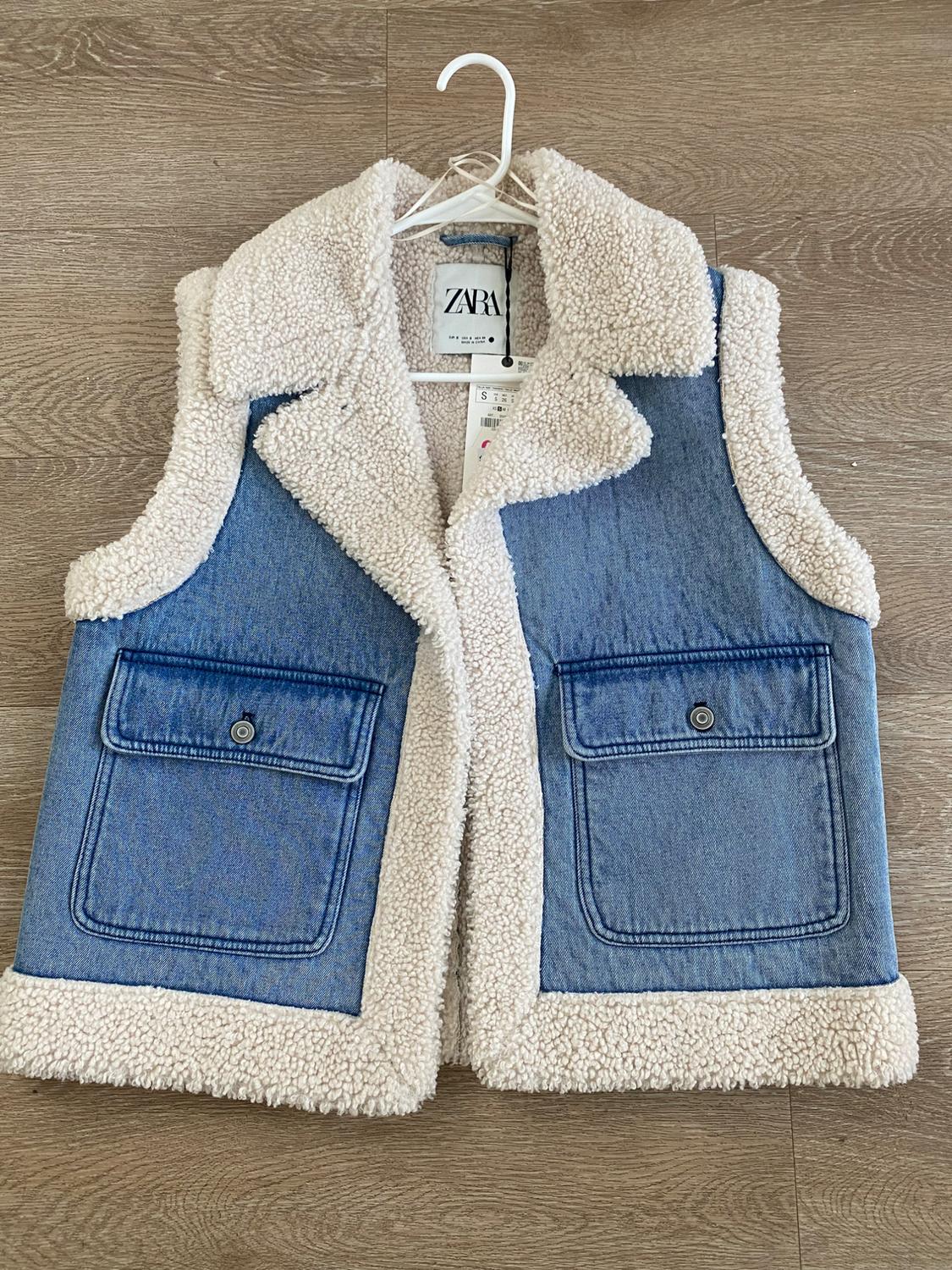 Denim vest with white shearling lining, two front pockets, and visible brand tag hanging on a white hanger.