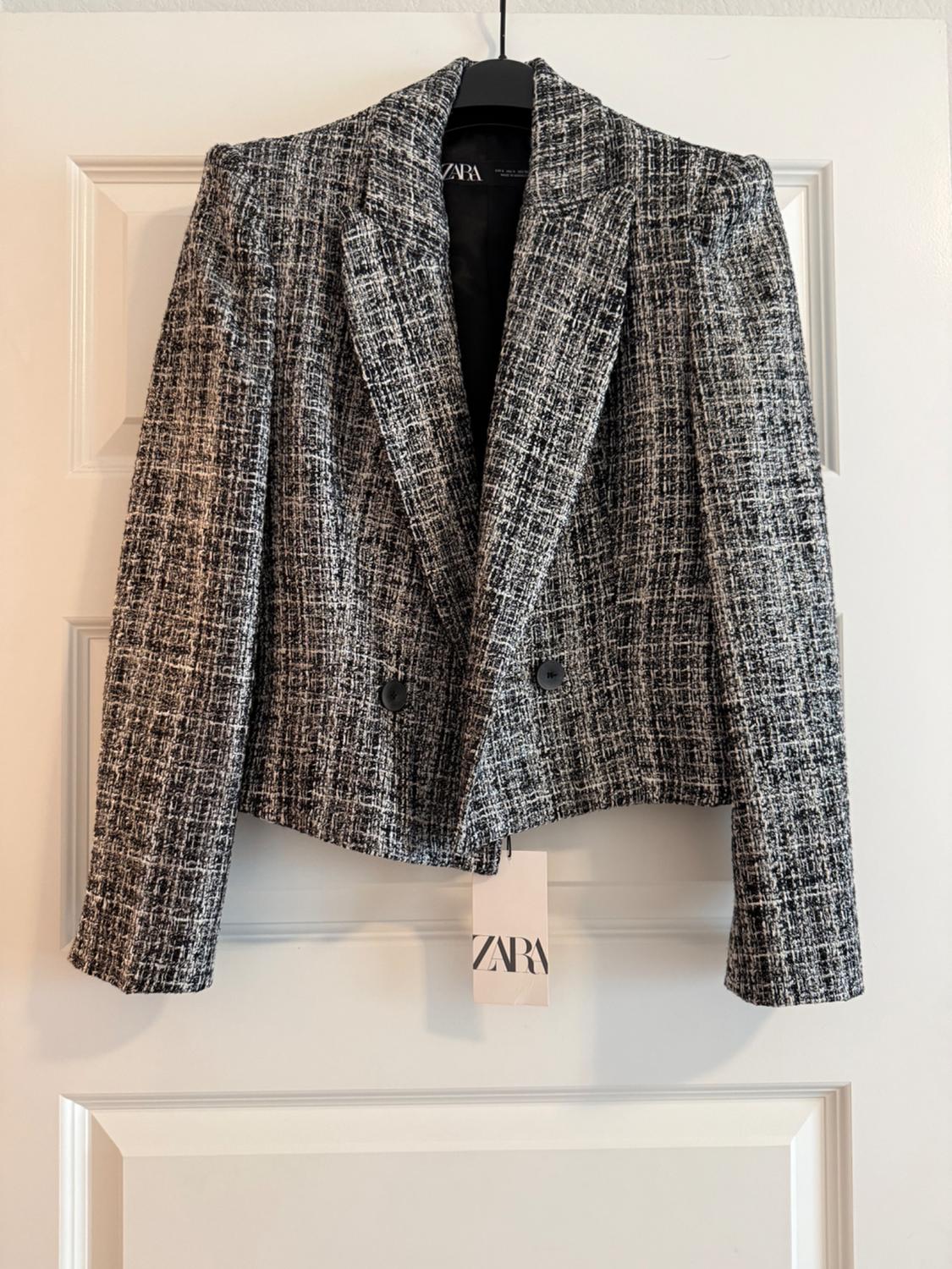 Textured black and white blazer hanging on a hanger on a white door. Buttons and labels with the text ZARA are visible.