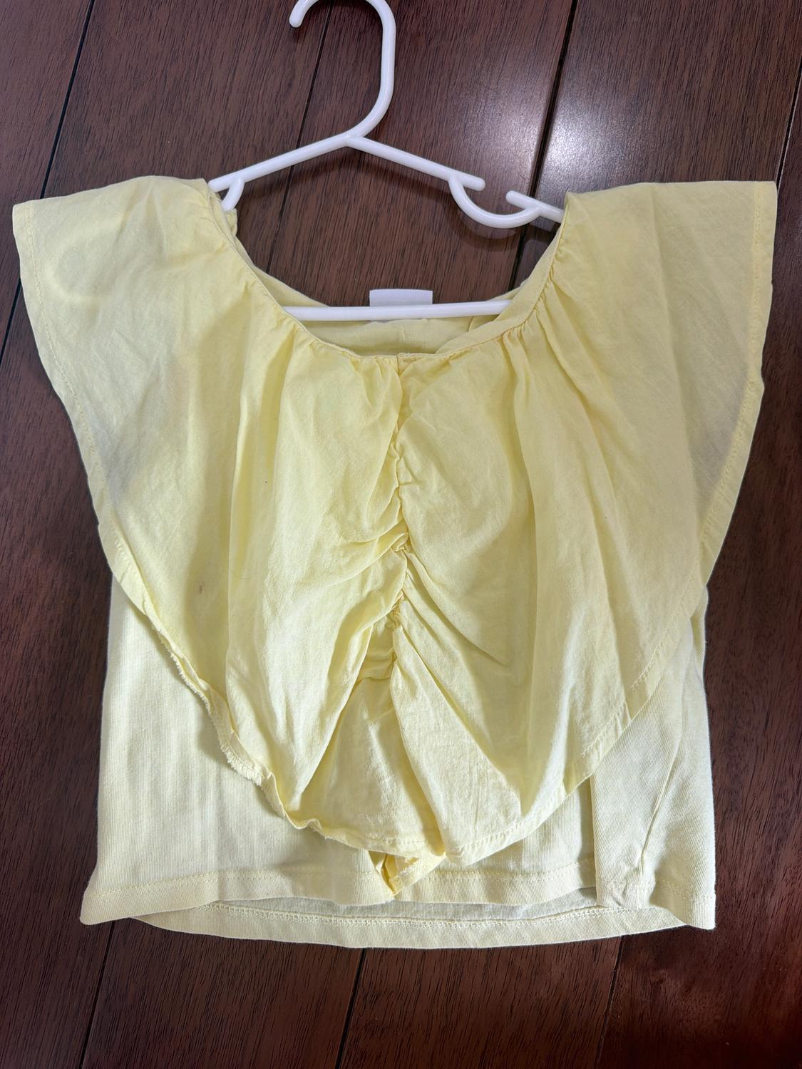 Yellow short-sleeved blouse with ruffles, hanging on a white hanger over a brown wooden floor.