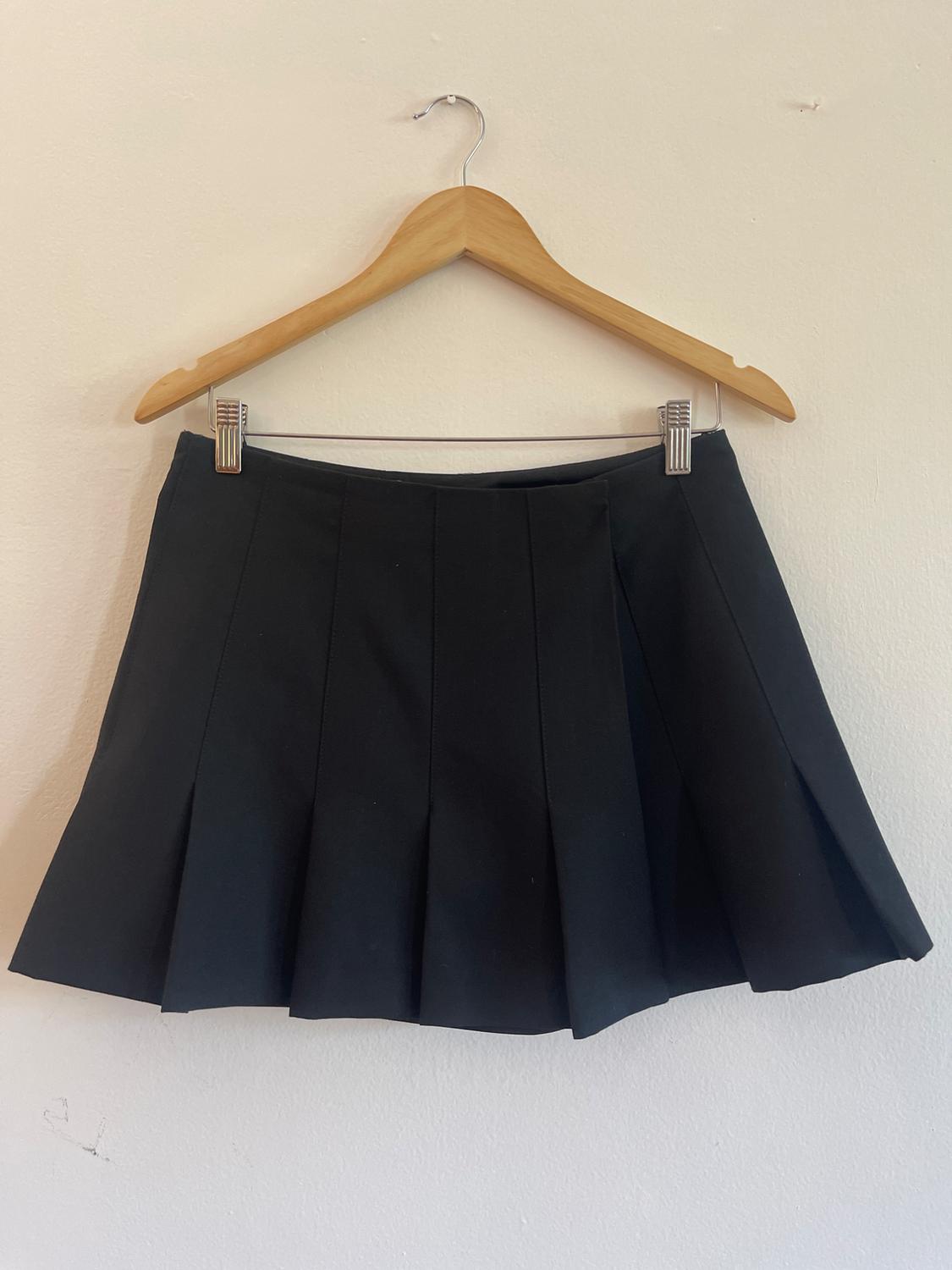 Black pleated miniskirt hanging on a wooden hanger with clips against a light-colored wall. Simple and casual composition.