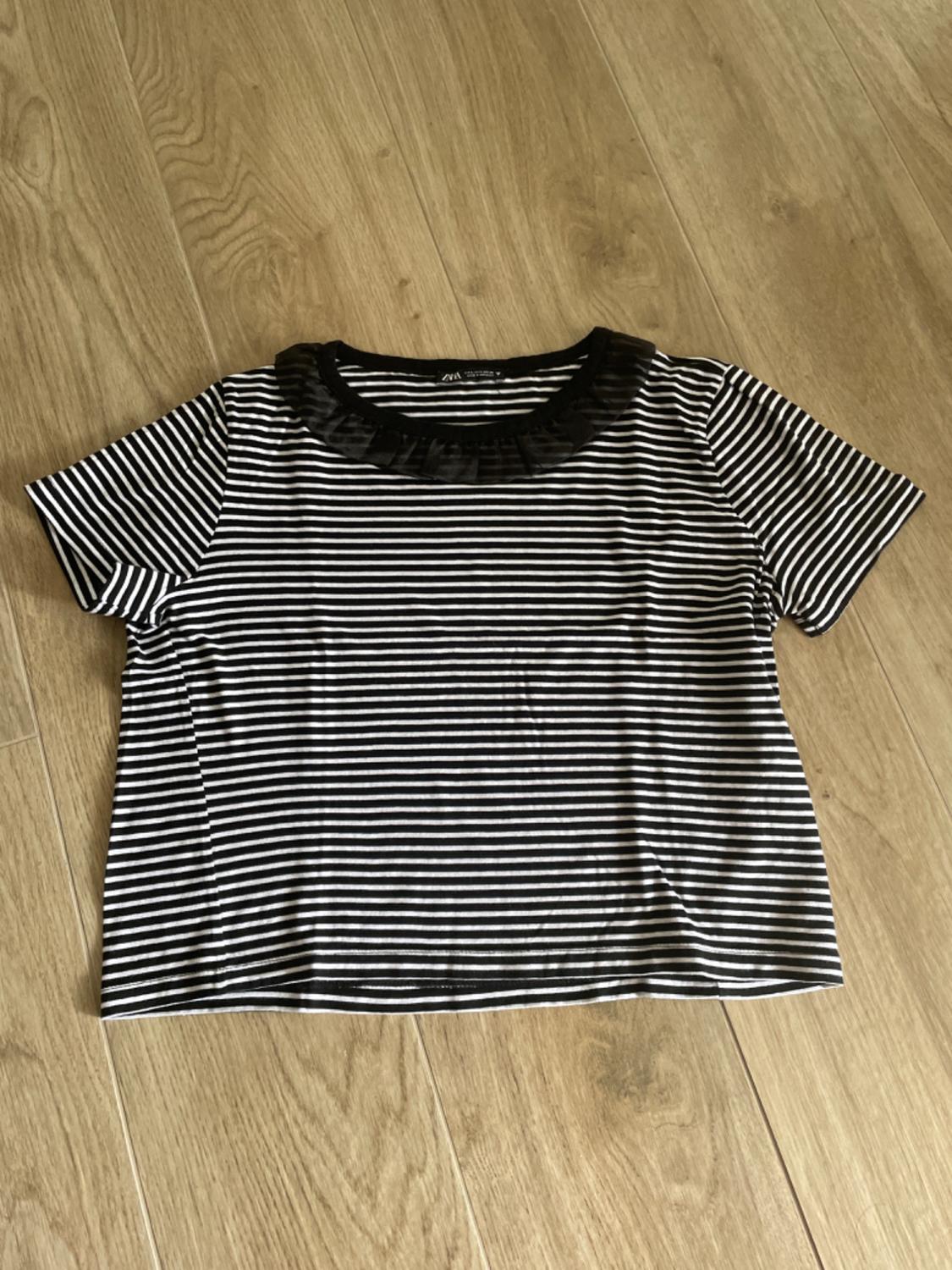 Black and white striped t-shirt with black pleated collar, on light wooden floor. Label visible on the neck.