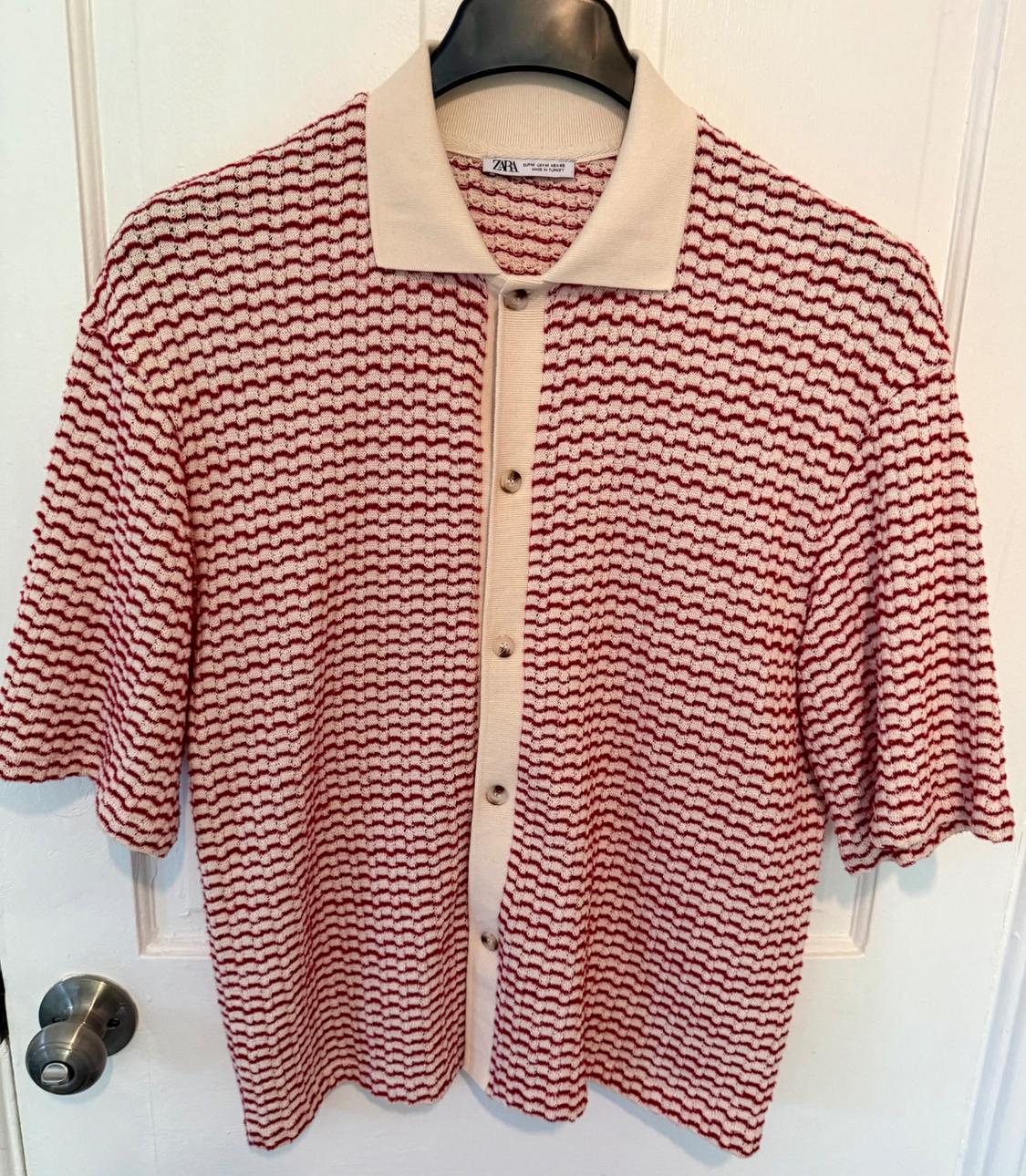 Knitted shirt with a red and white wave pattern, beige collar and buttons, hanging on a black coat hanger over a white door.