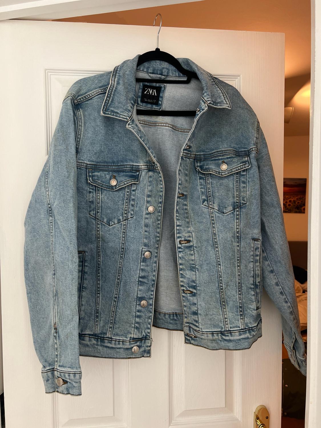 Blue denim jacket on a black hanger over a white door. Label with the text ZARA. Bedroom background, casual and relaxed atmosphere.