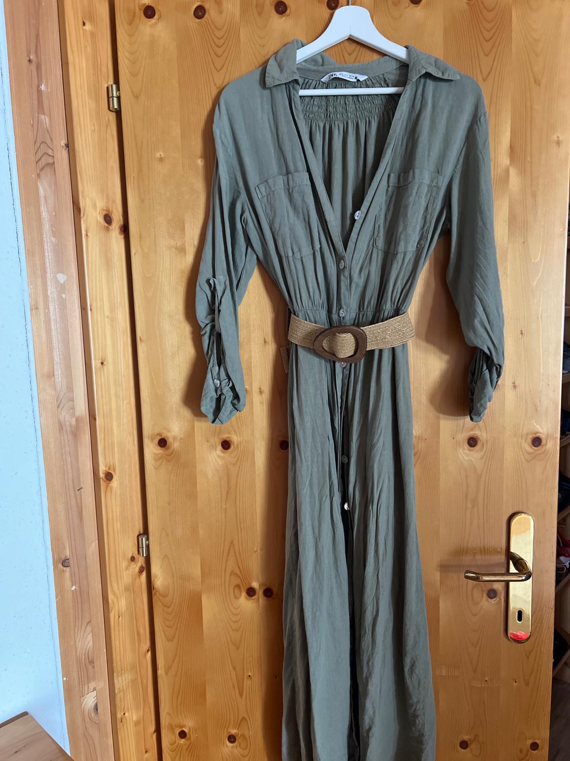 Olive green shirt dress with a braided belt, hanging from a hanger on a wooden door. ZARA label on the collar.