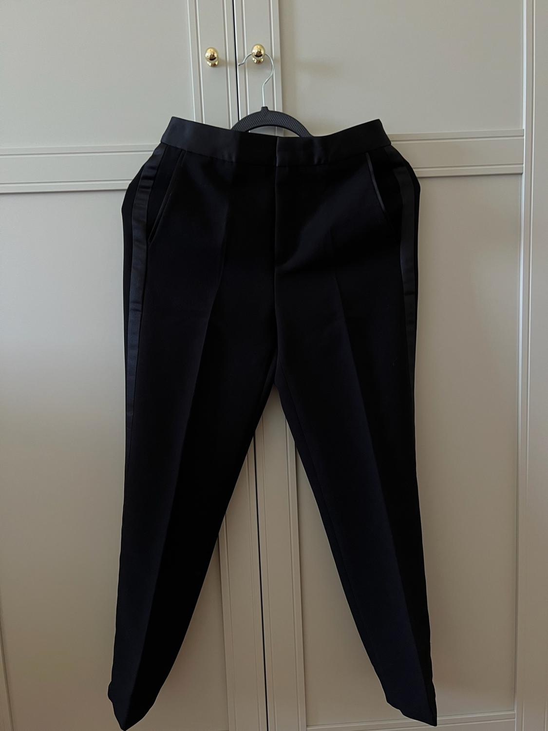 Black trousers with a satin side stripe, hanging on a hanger in front of a white wardrobe with gold knobs.