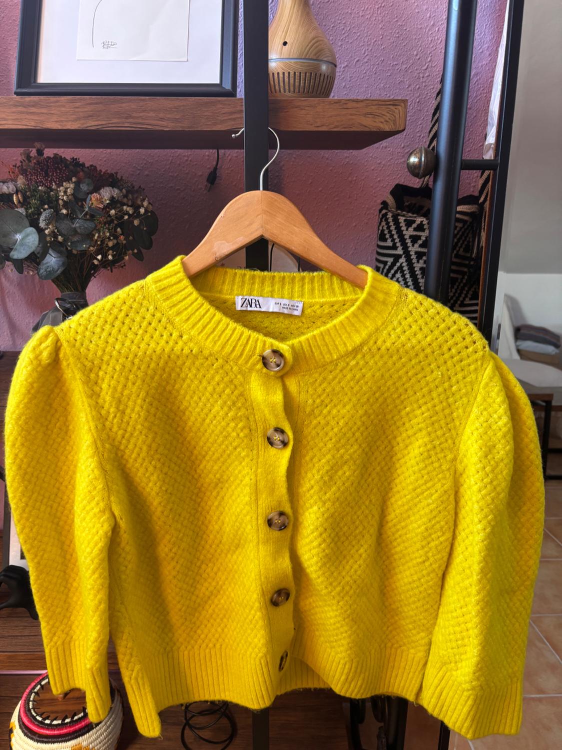 Yellow cardigan with wooden buttons hanging on a hanger. Background with shelving, a vase of dried flowers and decor.