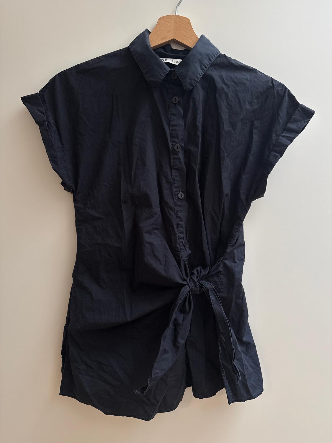 Black short-sleeved blouse with buttons and a tie at the waist, hanging on a wooden hanger against a light background.