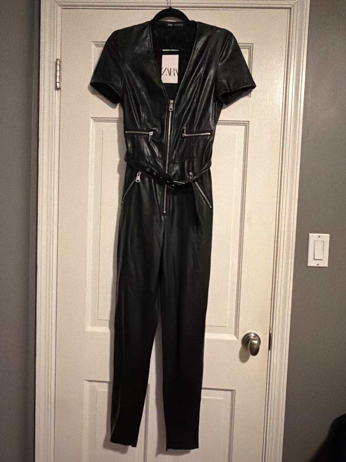 Black short-sleeved jumpsuit with zips and a belt, hanging on a white door. Label with the text ZARA.