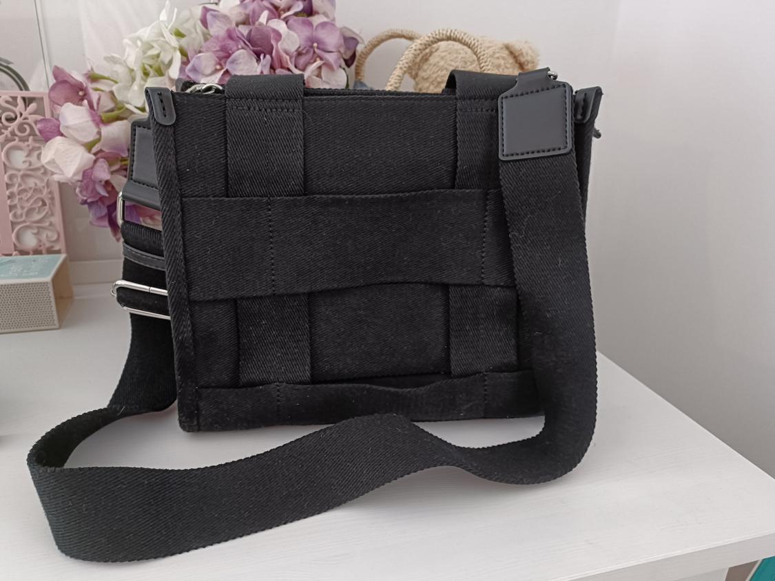 Black cross-body bag with a woven design and wide fabric strap, resting on a white surface with purple flowers in the background.