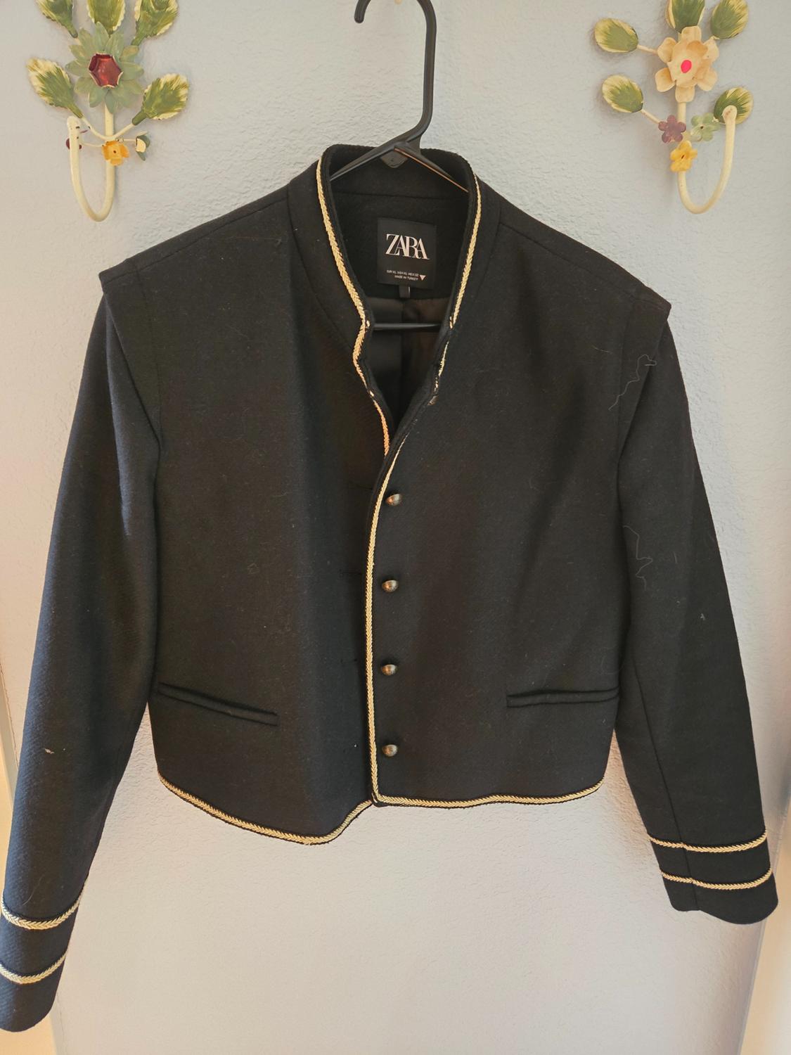 Black jacket with gold trim hanging on a hanger. Background with floral decoration on the wall. Visible label: ZARA.