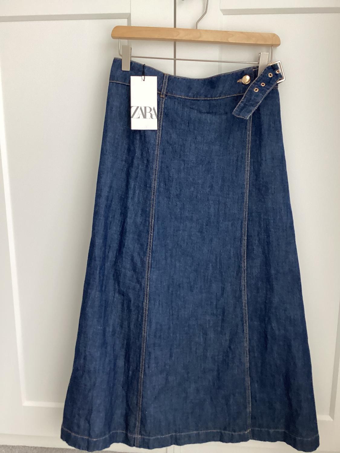 Long blue denim skirt with belt and ZARA tag, hanging on a wooden hanger in front of a white door.