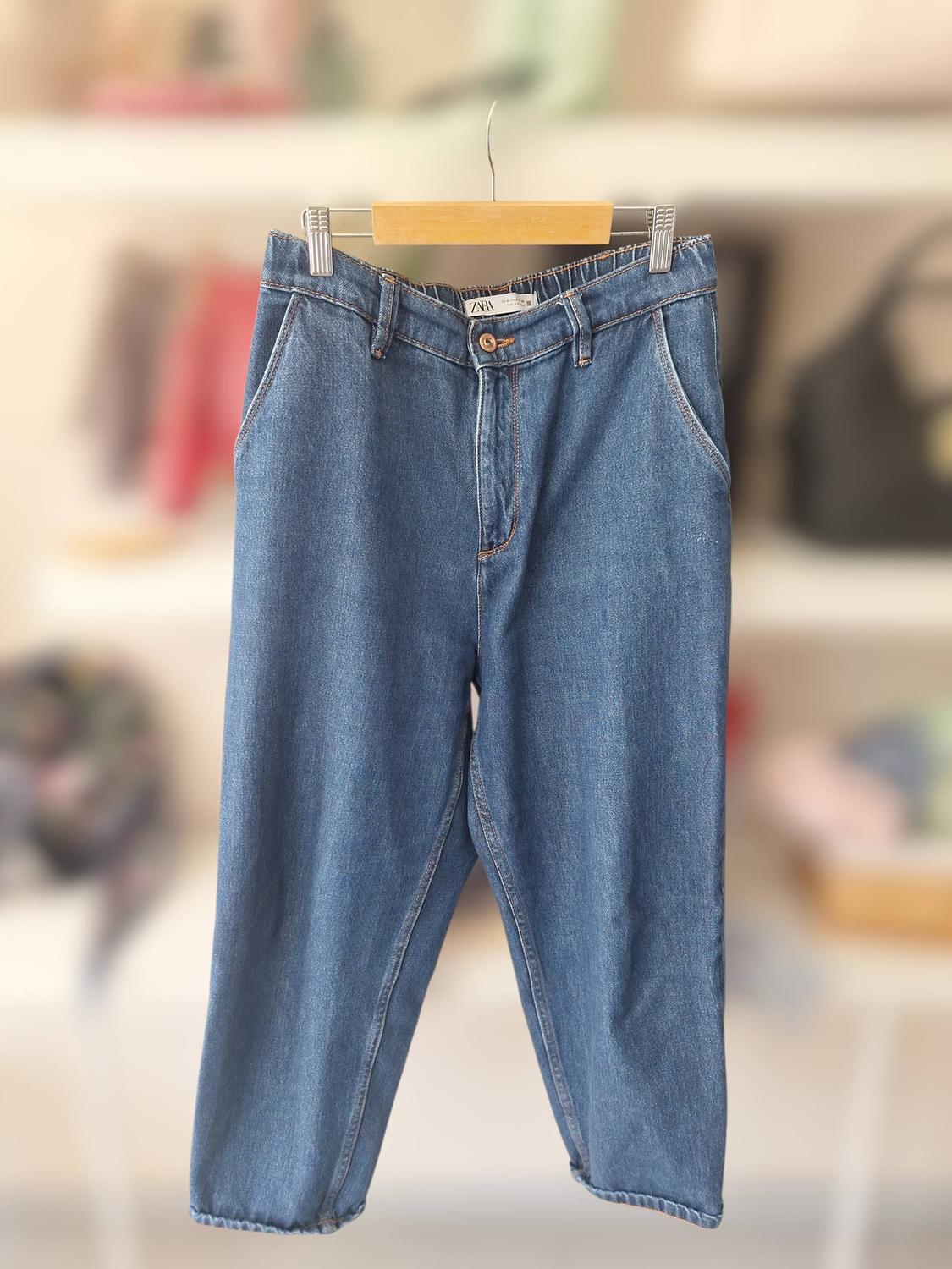 Blue denim jeans hanging on a wooden hanger, blurred background with shelves and handbags. Visible label: Zara.