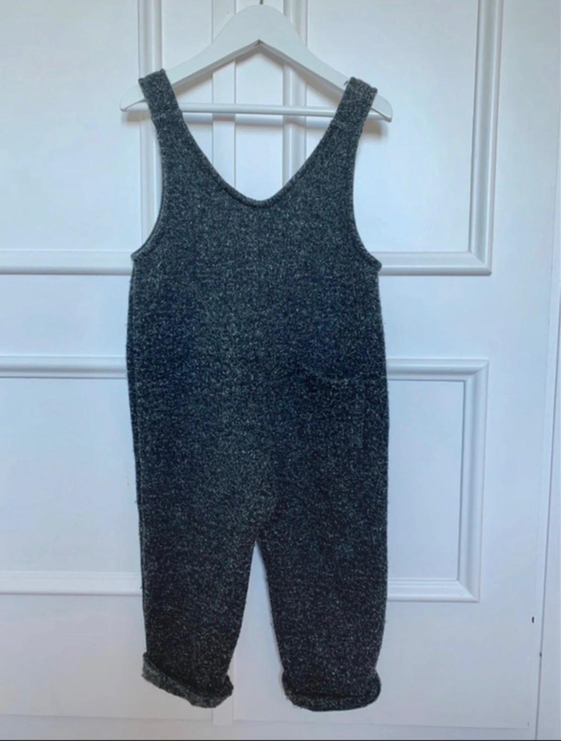 Grey jumpsuit with thick straps hanging on a white hanger over a white paneled door.