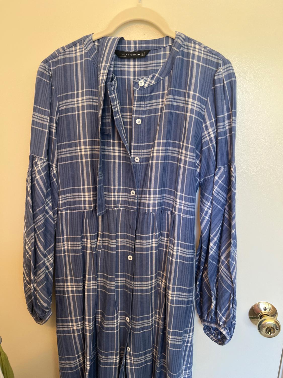 Blue and white checked dress with buttons and puff sleeves, hanging on a door. Label with text ZARA WOMAN.