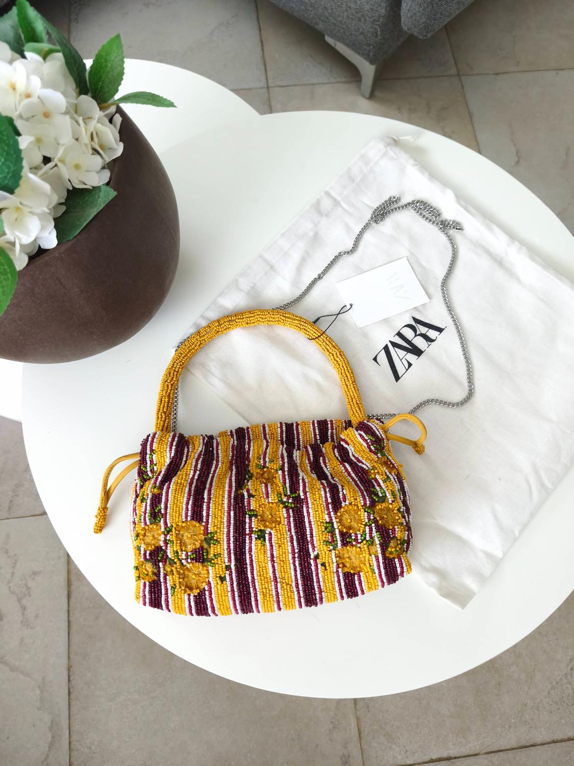 Striped and floral beaded bag with chain, on a bag with the text ZARA on a white table next to a pot with flowers.