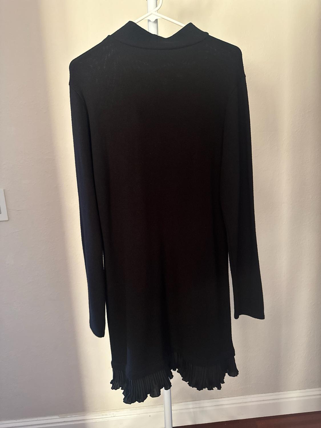 Long-sleeved black high-neck dress with pleated ruffle detailing on the hem, hanging on a white hanger in front of a wall.