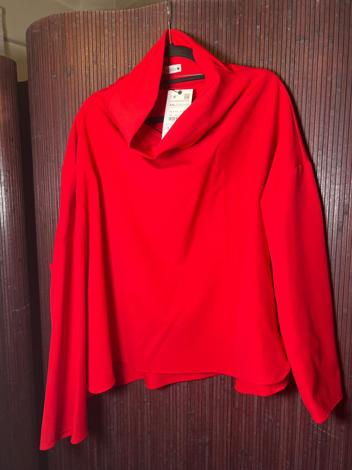 Red long-sleeved blouse with a wide collar, hanging on a hanger in front of a brown wood-paneled background. Visible tag.