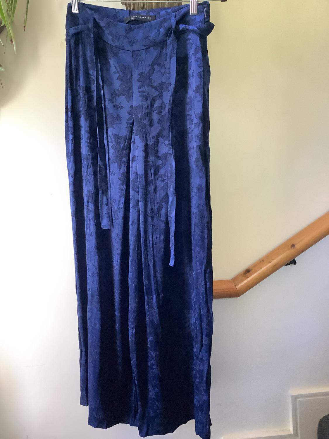 Dark blue long trousers with a floral print, hanging on a hanger, with a light background and a visible wooden railing.