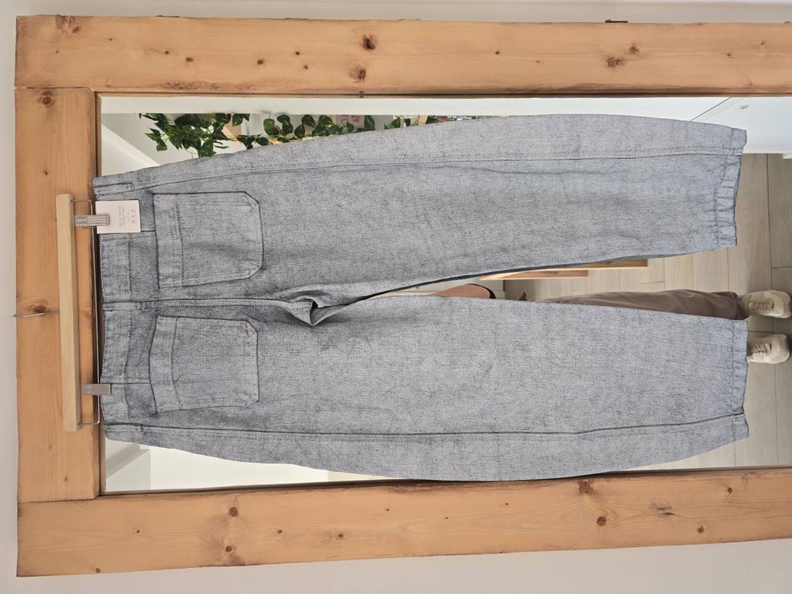Grey trousers hanging on a wooden frame, with visible back pockets. Background with plants and light wooden floor.