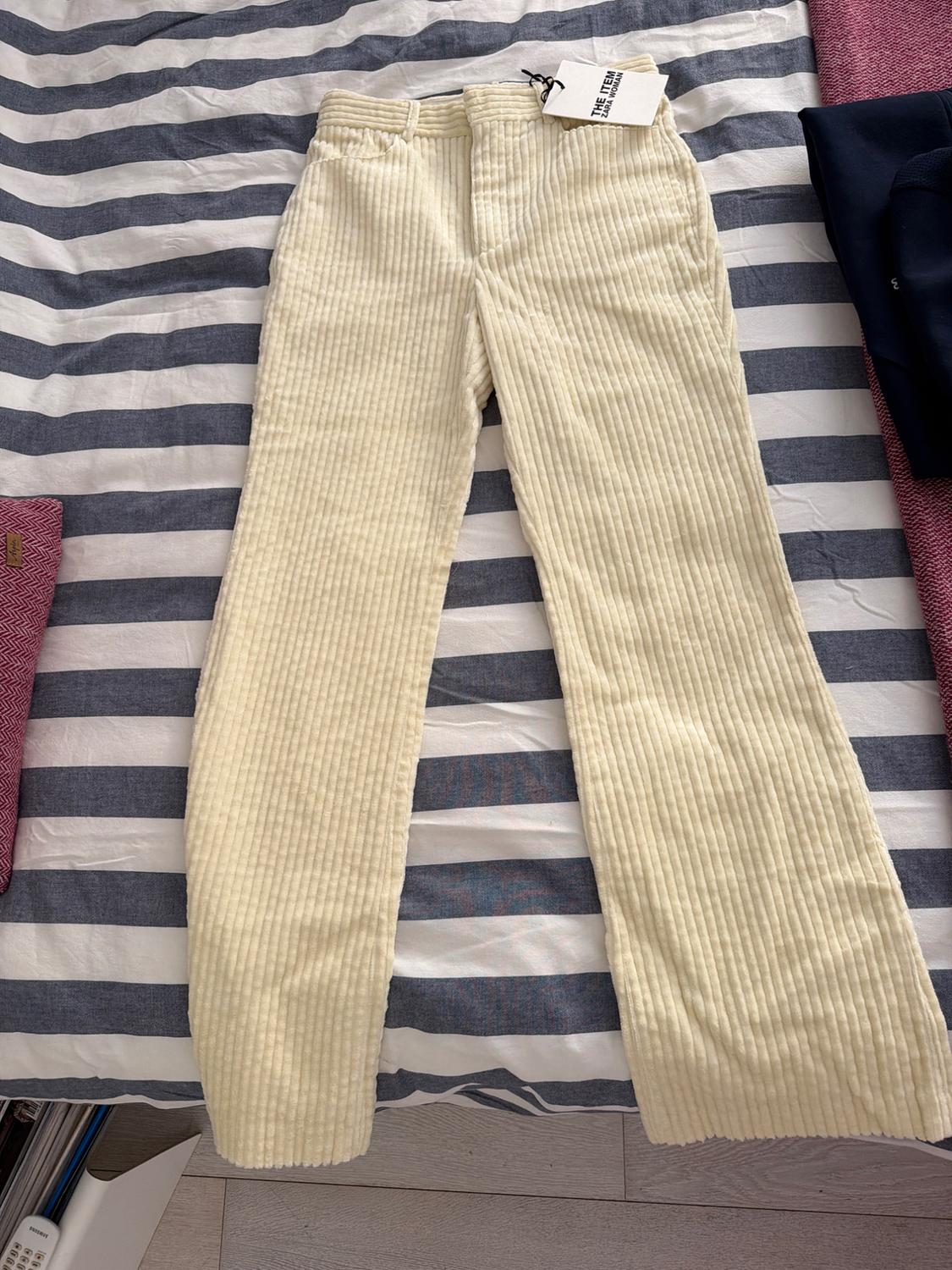 Beige corduroy trousers with a 'The New' label on a bed with a white and grey striped quilt.