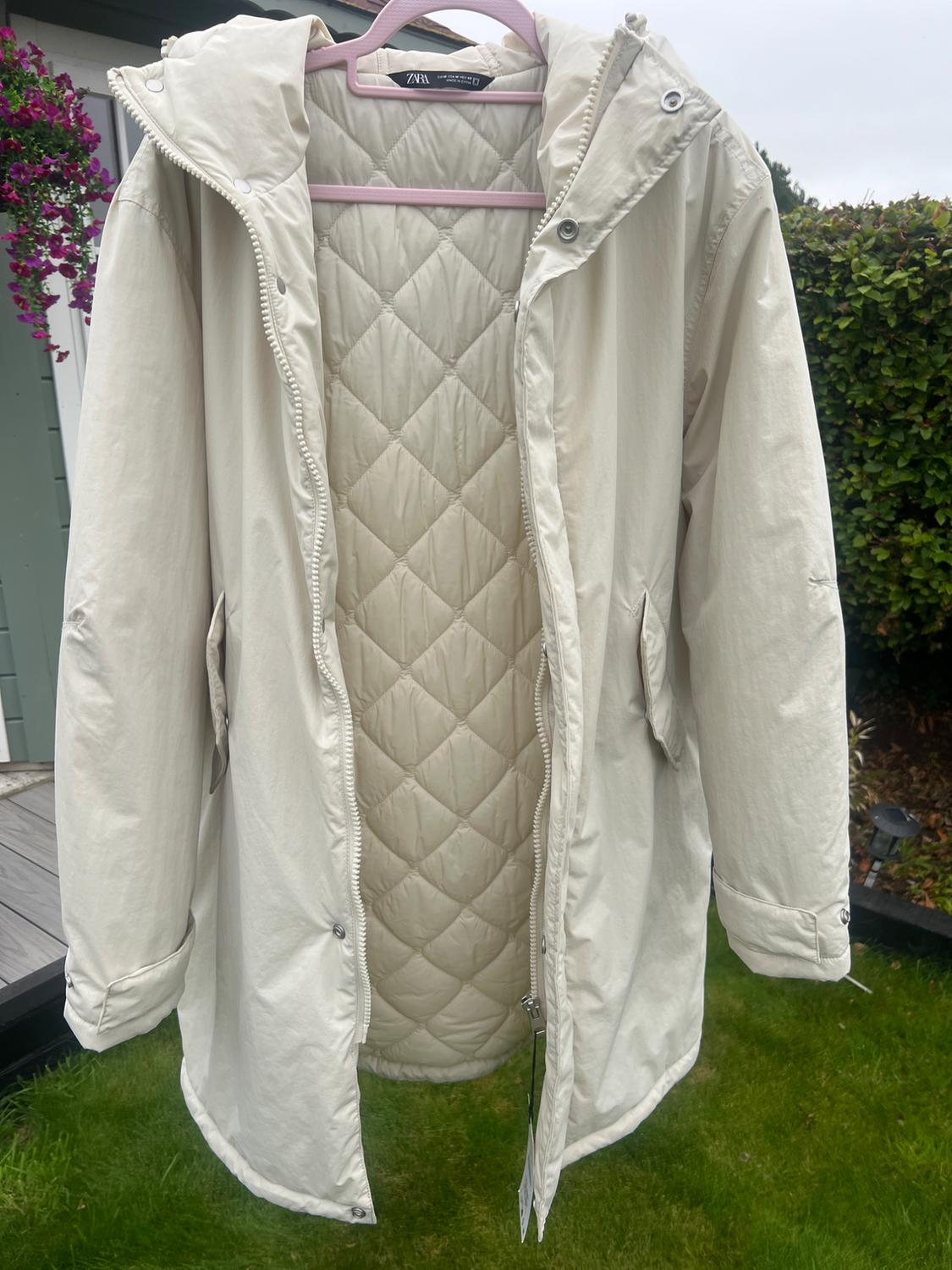Beige hooded coat hanging from a pink clothes hanger outdoors. It shows a quilted inner lining and the ZARA label on the collar.