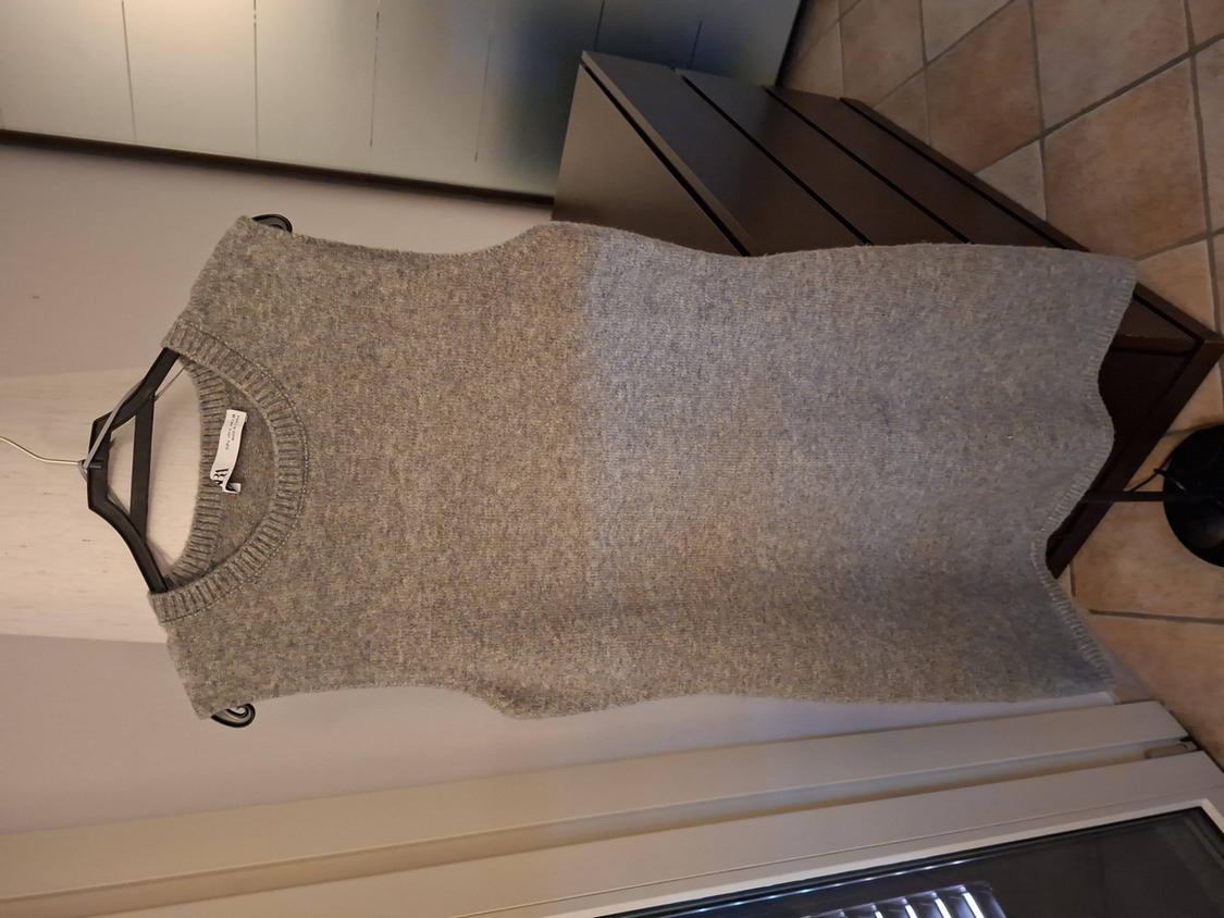 Grey knitted waistcoat hanging on a black hanger against a background of furniture and a tiled floor. Label visible on the collar.
