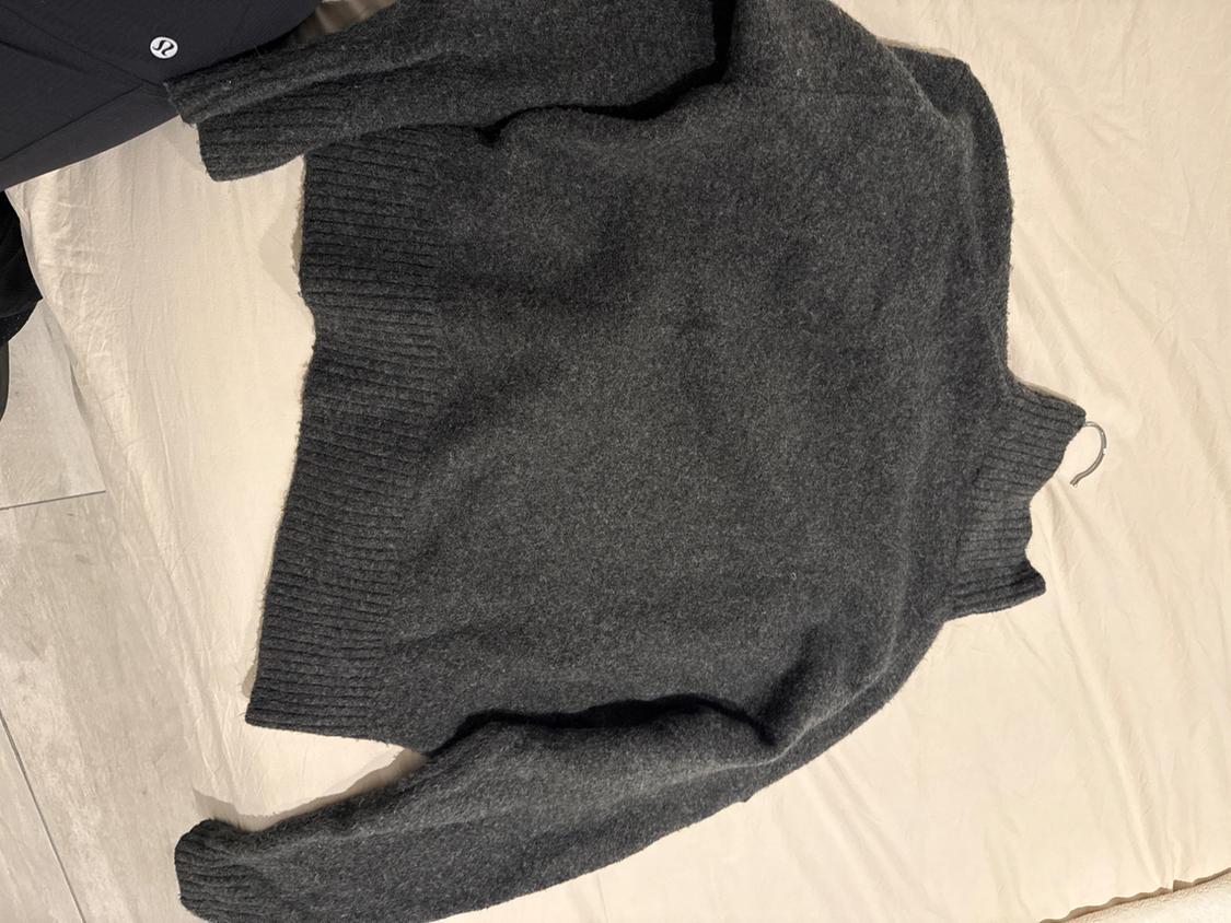 Dark grey chunky knit jumper on a beige bed. Part of a leg in black trousers visible in the top left corner.