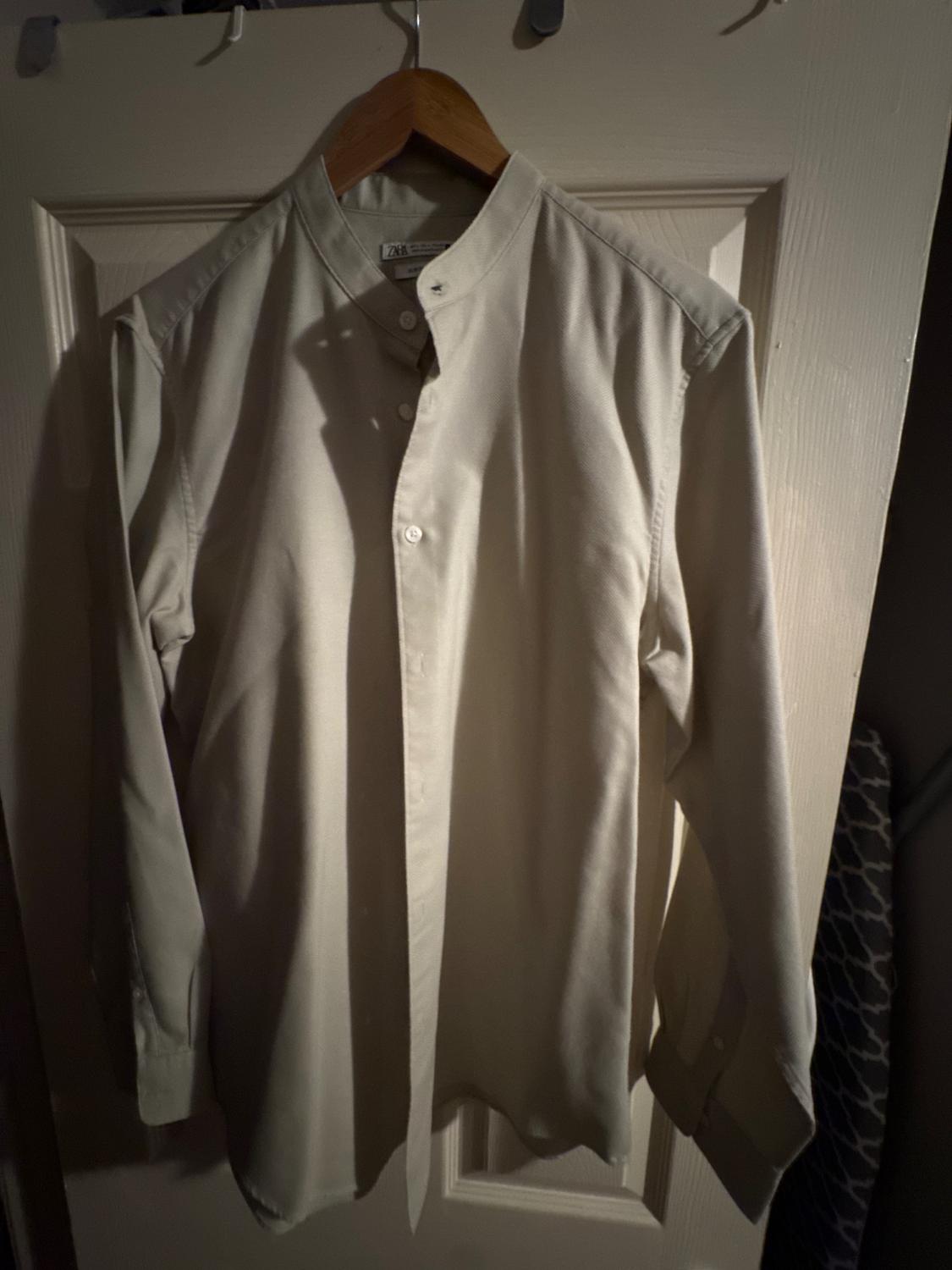 Beige grandad collar shirt hanging on a wooden hanger over a white door, illuminated with dim light.