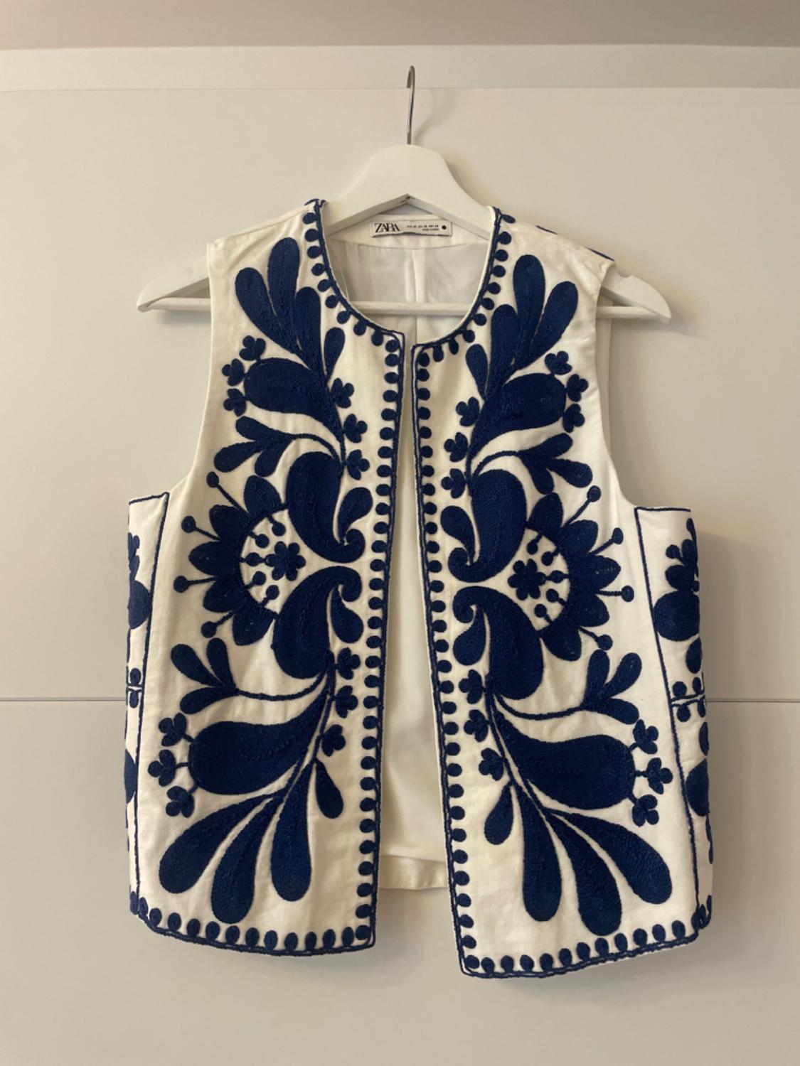 White waistcoat with blue floral embroidery, hanging on a white hanger. Visible label: "Zara Woman".