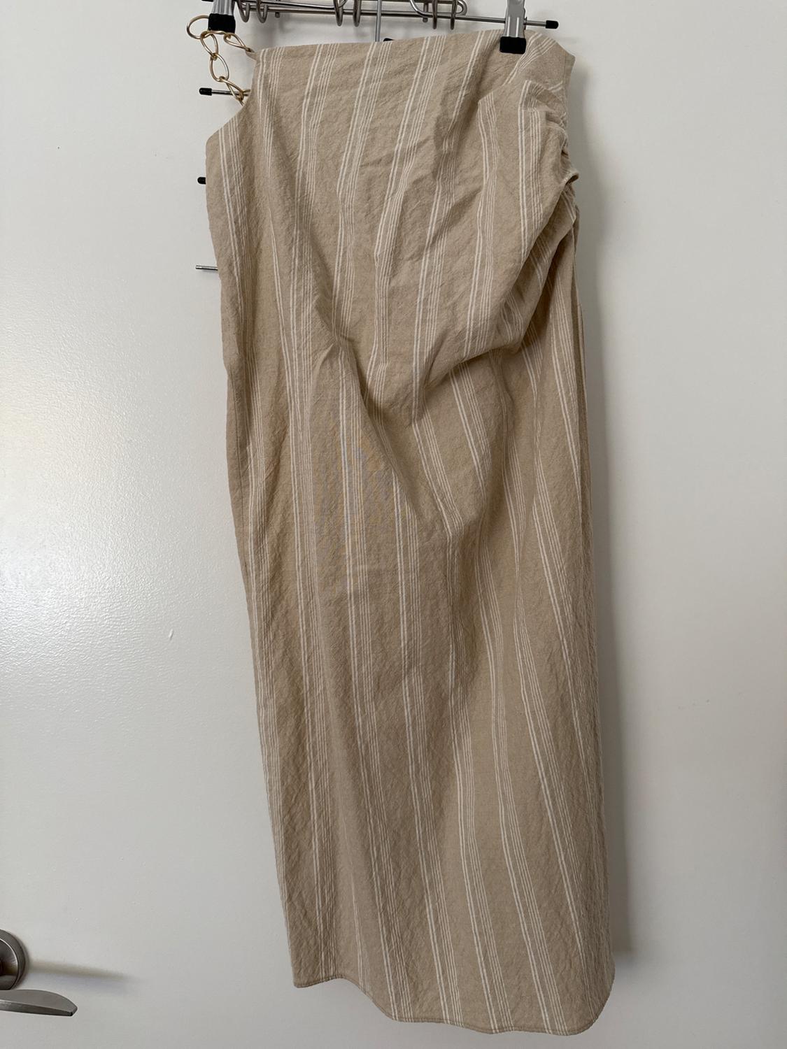 Beige skirt with vertical white stripes hanging on a hanger on a white door. It has a gold chain detail.