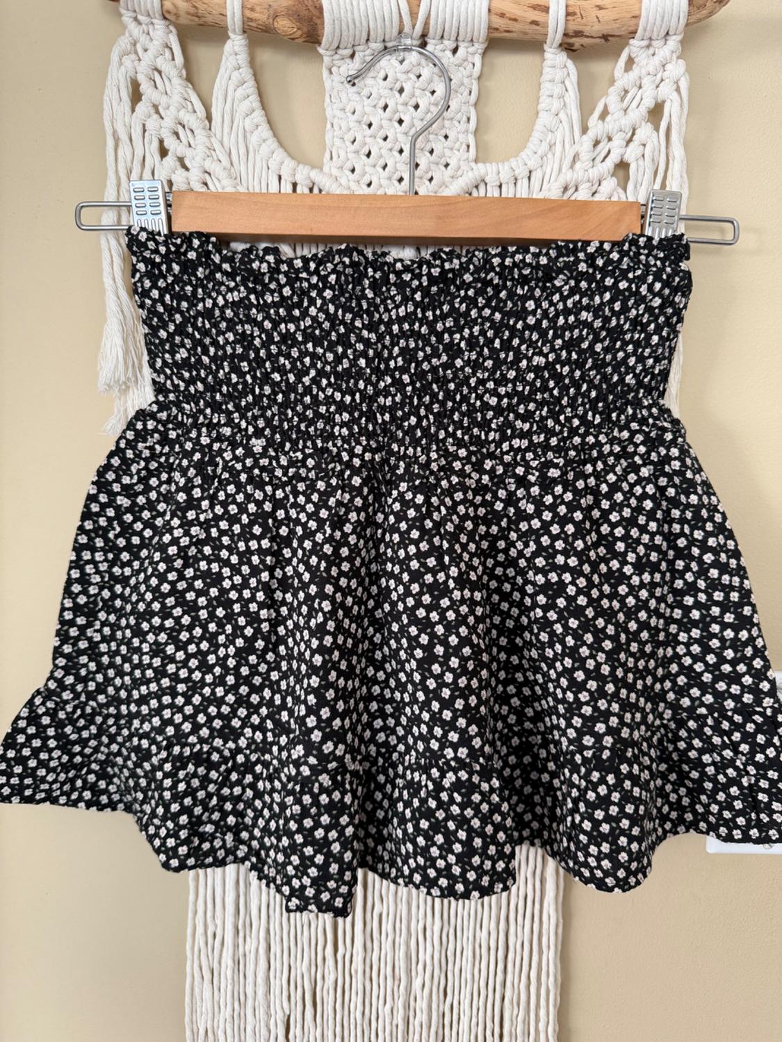 Black skirt with white floral print hanging on a wooden hanger against a white macramé background.