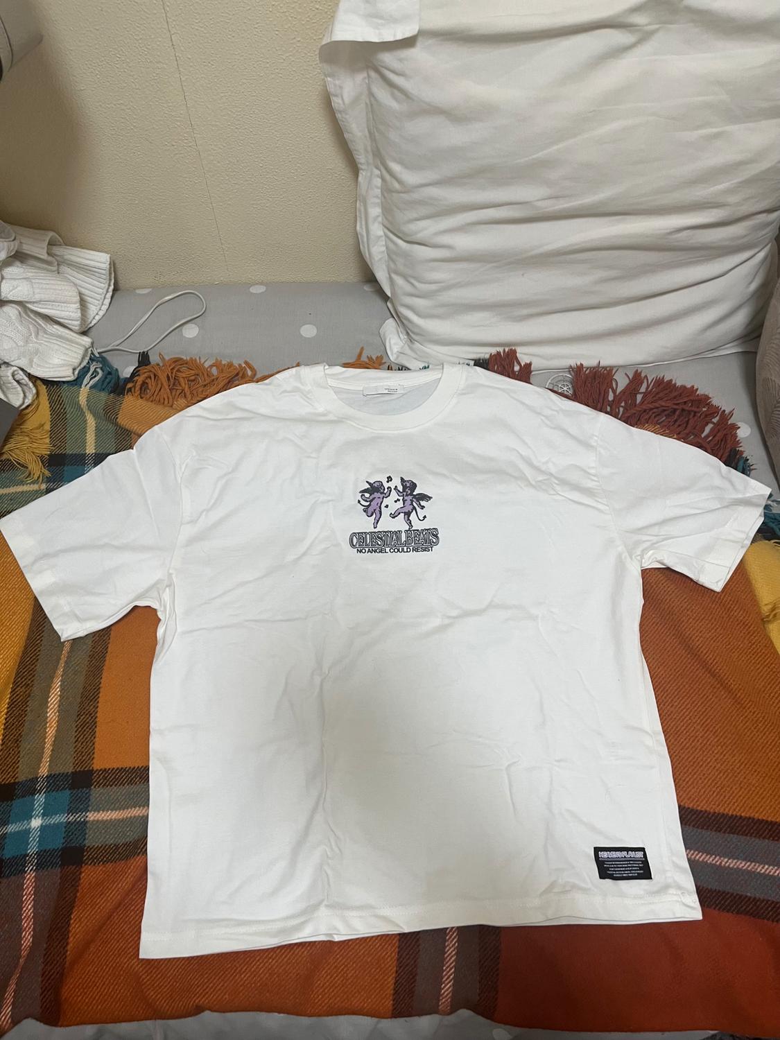 White t-shirt with a design of two figures in the centre and text "CELESTIAL BEINGS NO ANGEL COULD RESIST". Background with cushions and a checked blanket.