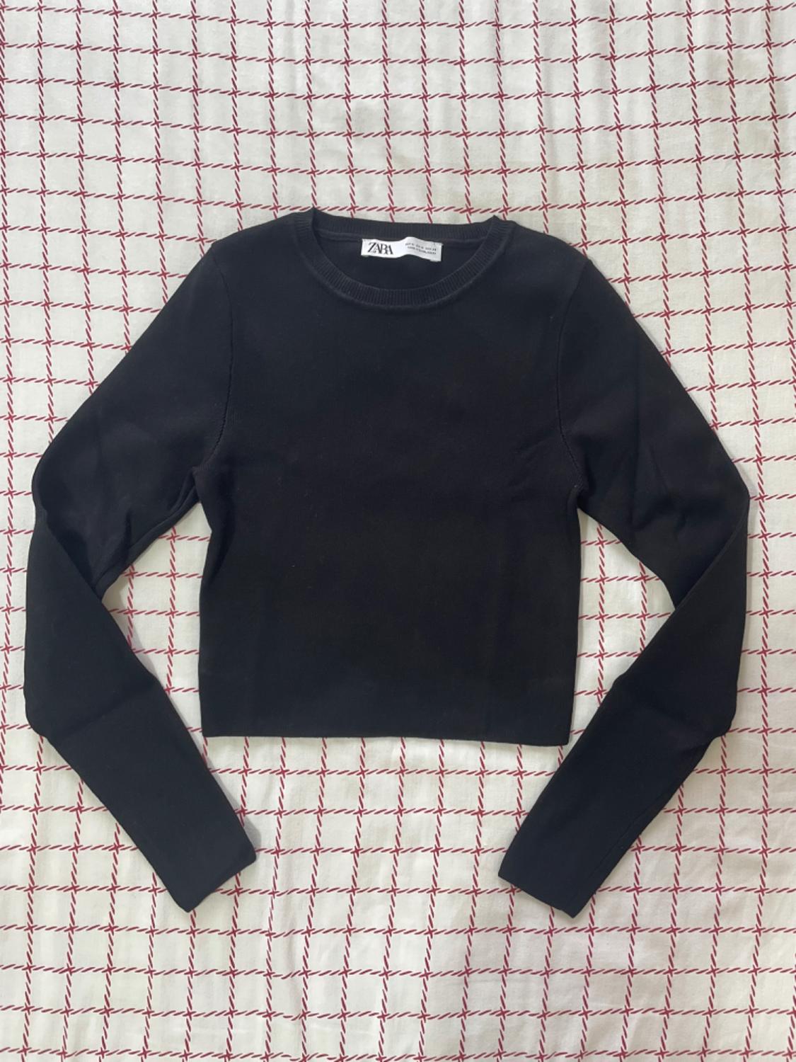Black long-sleeved jumper on white fabric with a red check pattern. Visible label with "Zara" text.