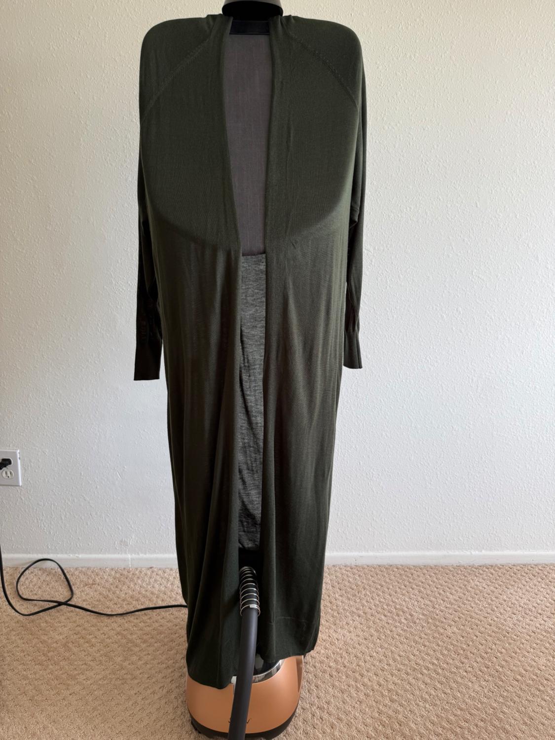 Mannequin in a dark green, long dress, open at the front, on a beige carpet. White wall background and visible cable on the floor.