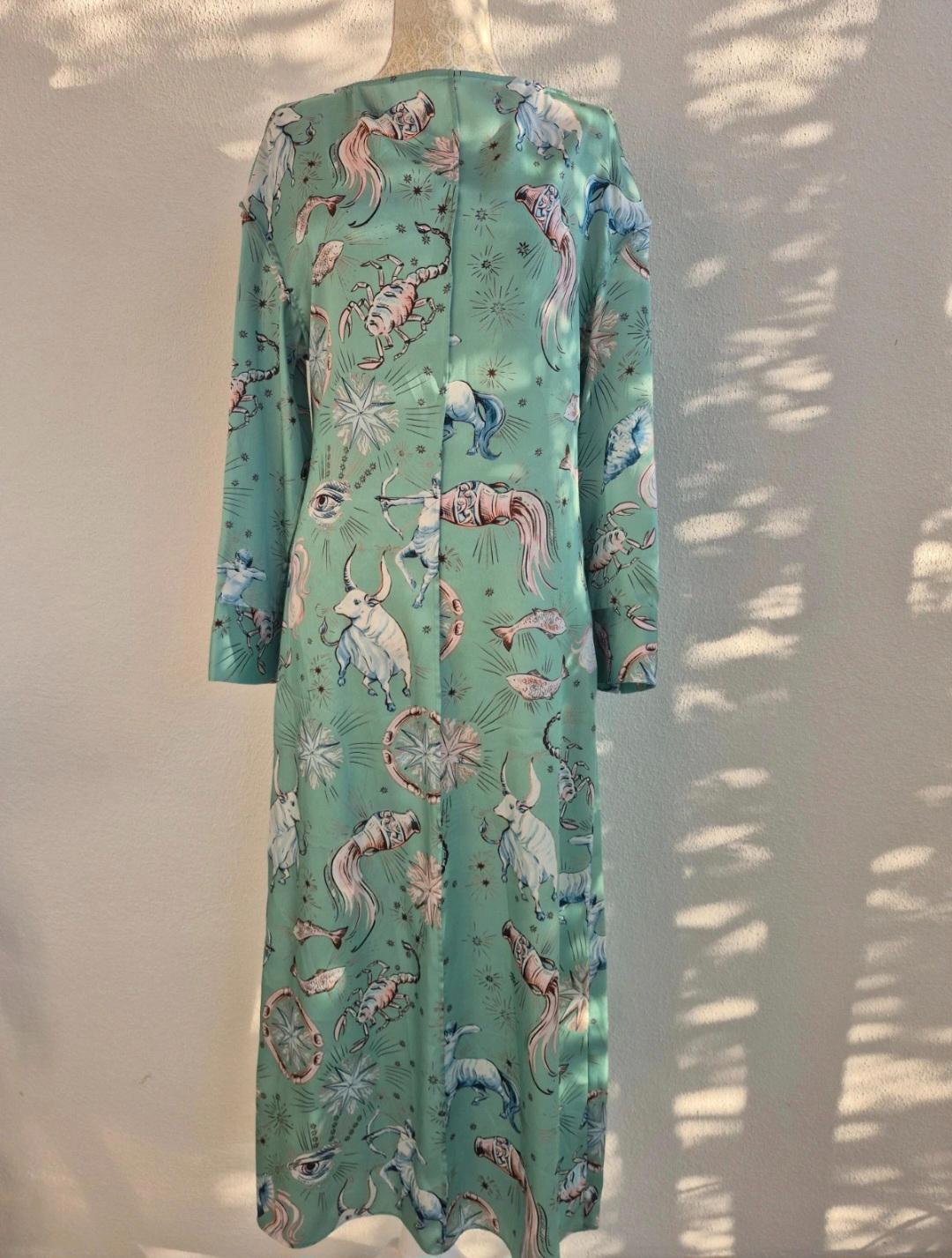 Long green dress with animal print and celestial figures. White background with projected leaf shadows.
