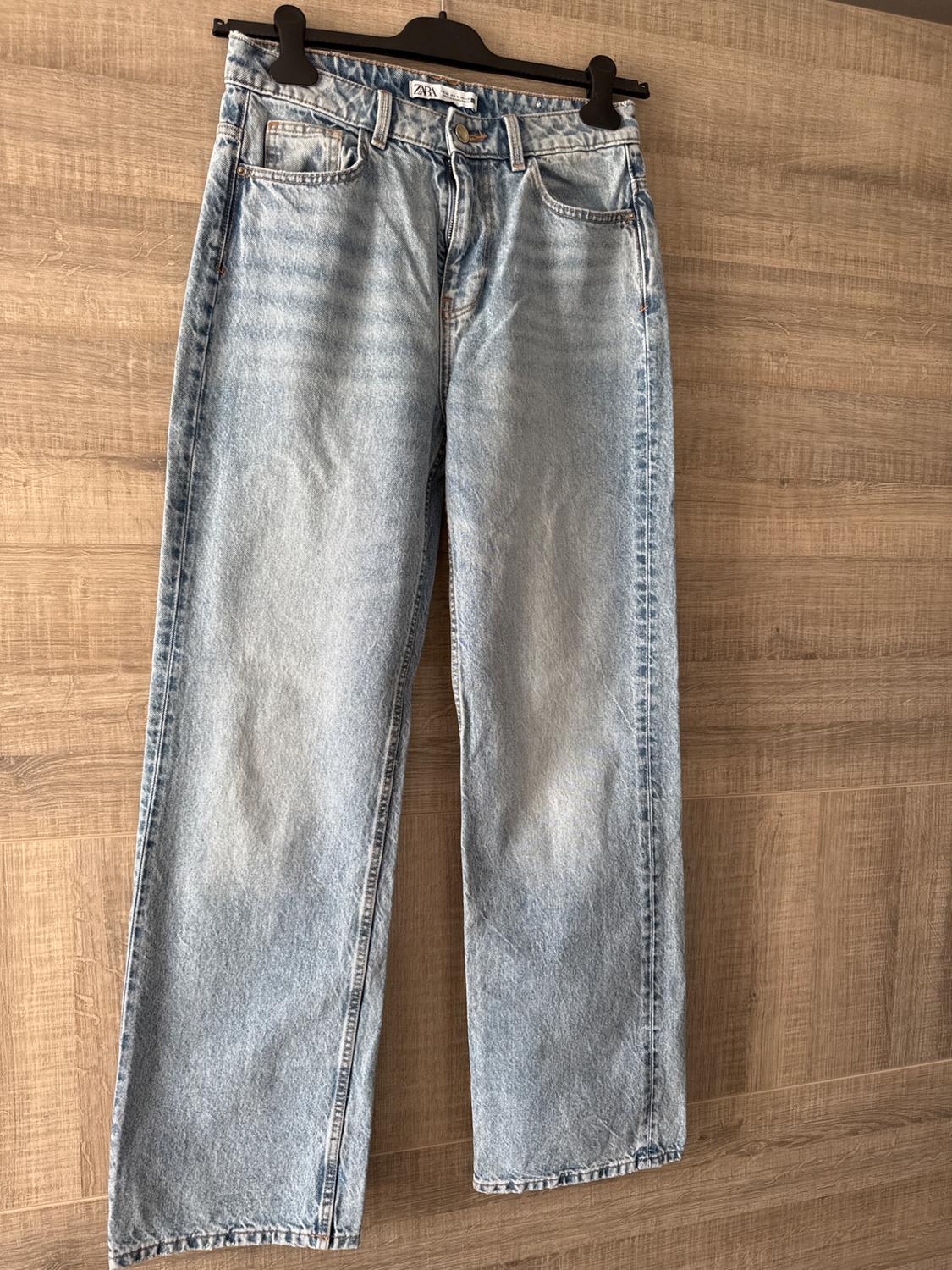 Light blue denim jeans hanging on a black hanger against a light wooden background. Label visible on the waistband.