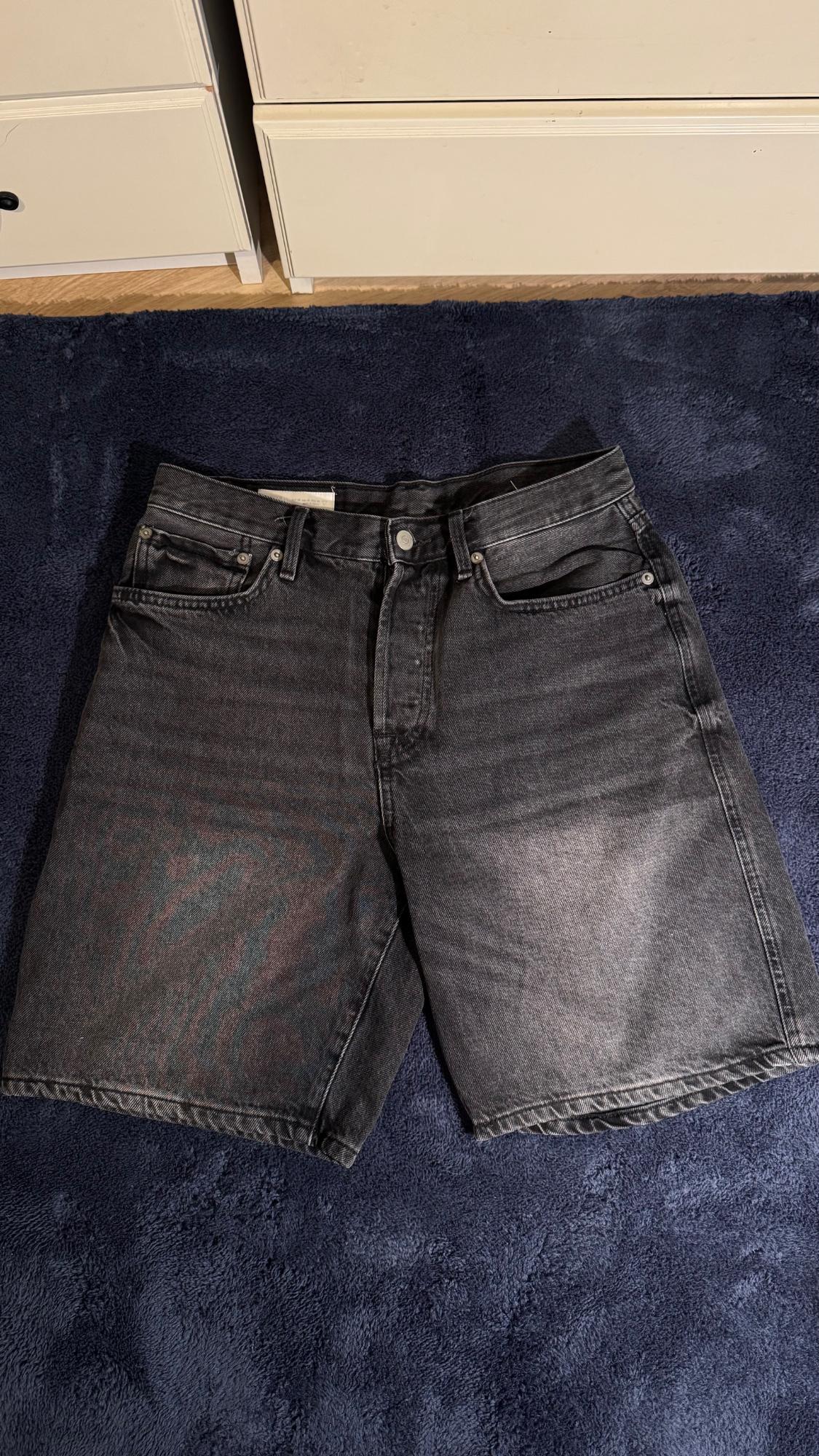 Black denim shorts with a washed effect, placed on a dark blue rug in front of a white piece of furniture.
