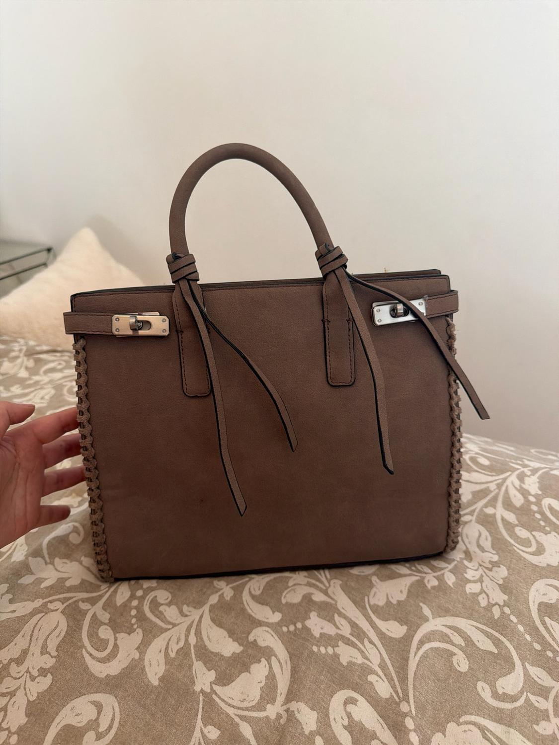 Brown handbag with handles, braided edges and silver clasps, resting on a bed with a patterned bedspread. A hand can be seen on the left.