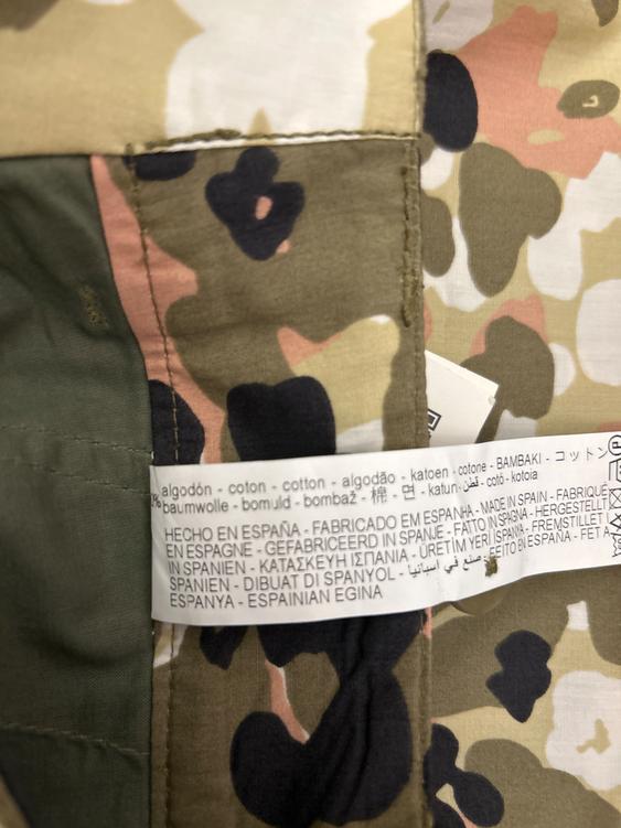 Camouflage fabric label in shades of green, beige, and pink. Visible text: "HECHO EN ESPAÑA - FABRICADO EN ESPAÑA - MADE IN SPAIN".